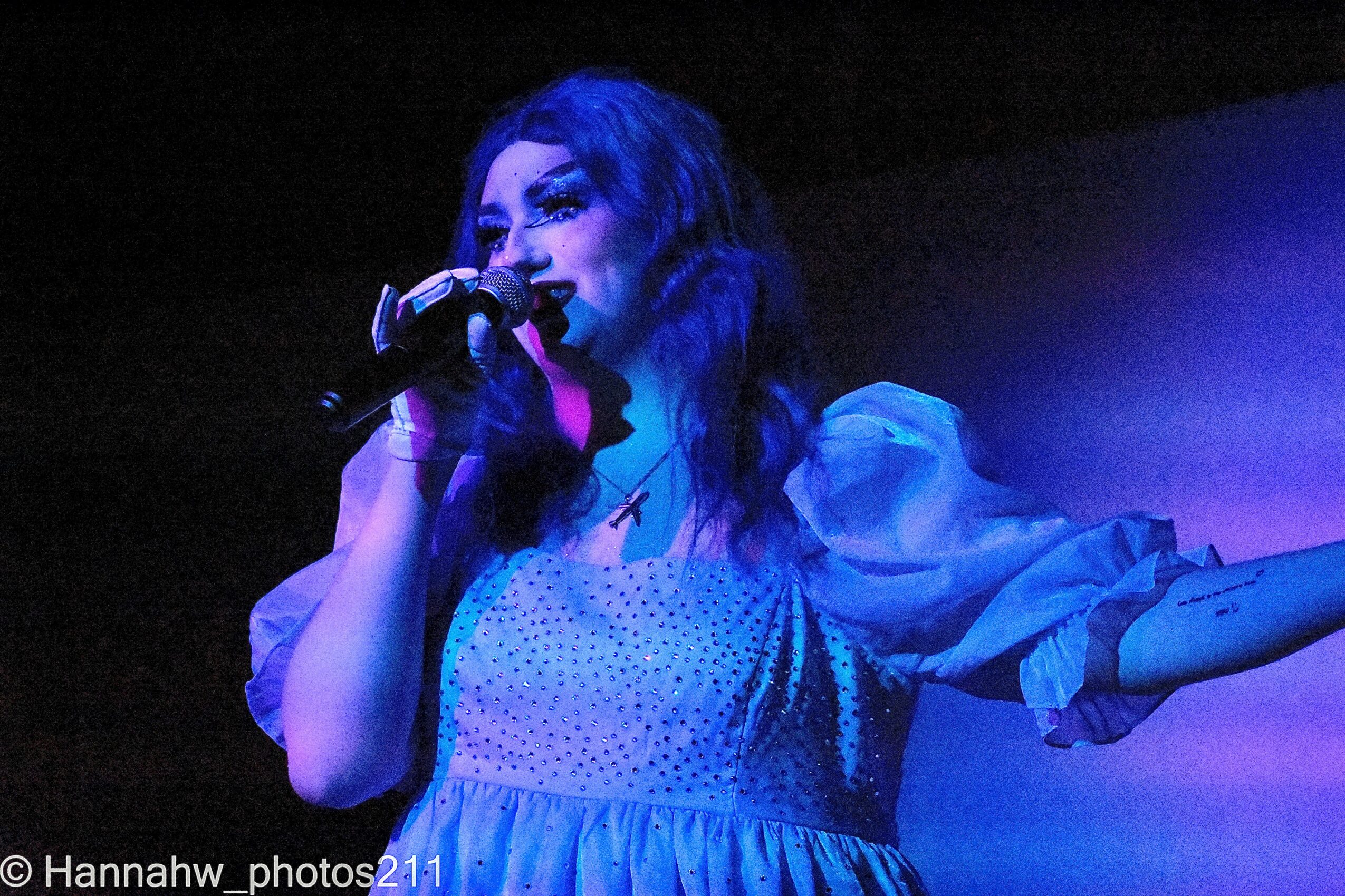 Person singing into a microphone on stage, with blue lighting and a dark background.