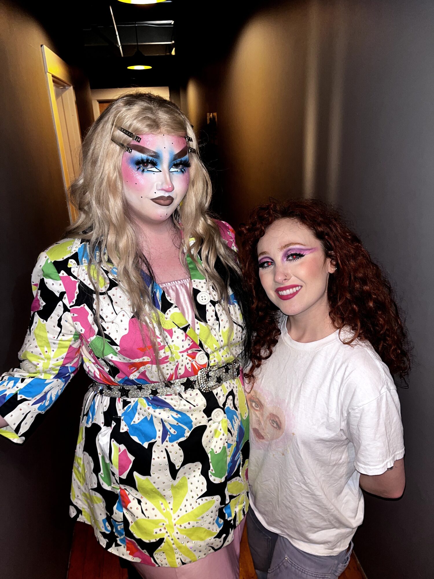 Two women with colorful makeup and curly hair standing in a hallway, one with elaborate face paint, smiling.