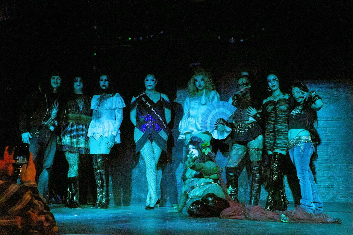 Group of nine women standing on stage, some holding fans, one sitting on the floor, in a dark setting with blue lighting.