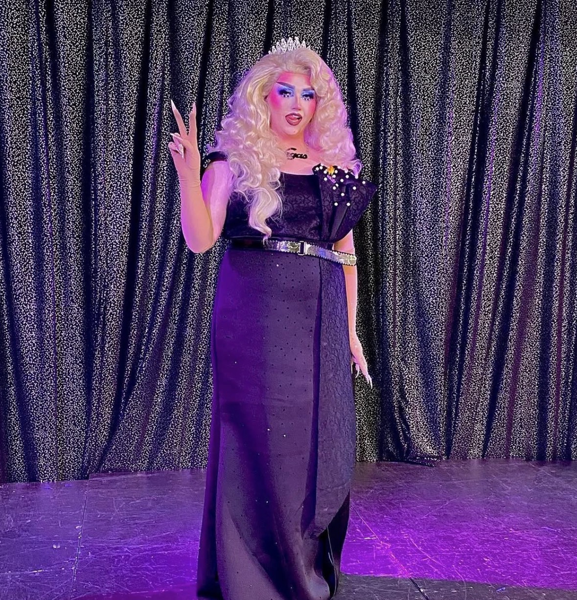Person with long curly blonde hair, wearing a black dress with shoulder embellishments, standing in front of a curtain, making a peace sign gesture.
