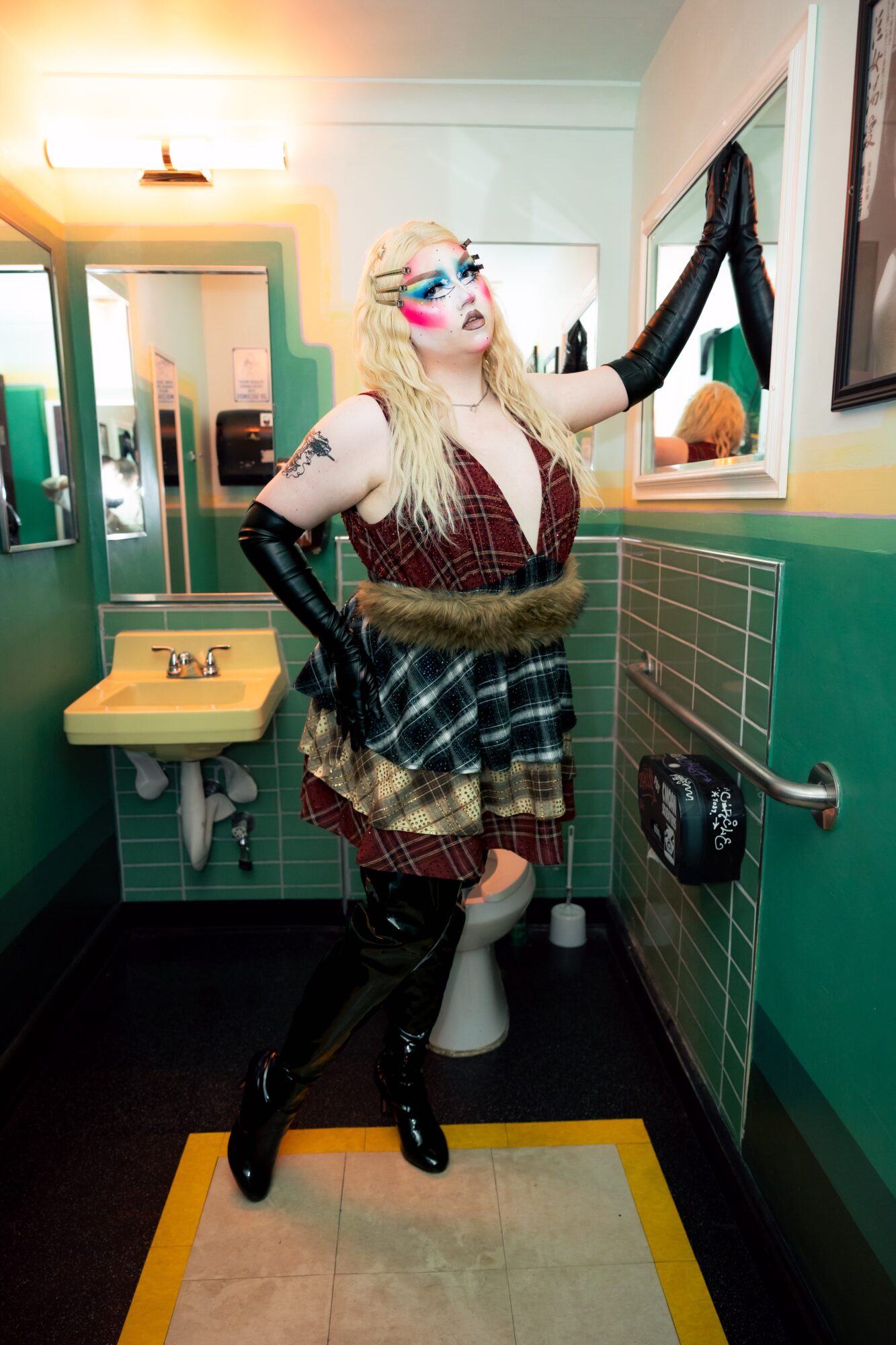 Person with long blonde hair, sunglasses, and colorful makeup, wearing a patterned dress and gloves, standing in a bathroom, looking at a mirror.