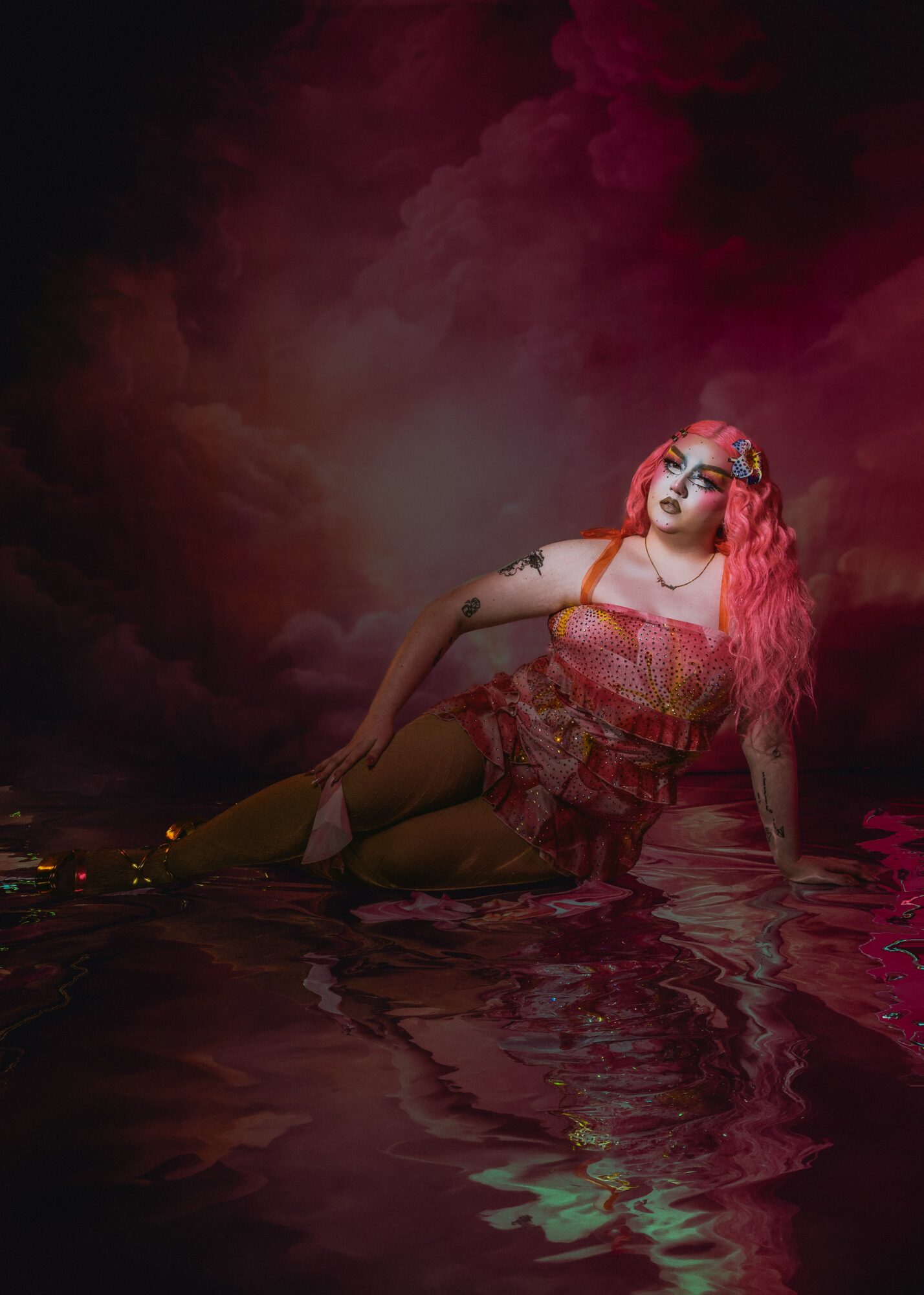 Person with pink hair in a colorful outfit, lying in water with dark, cloudy sky background.