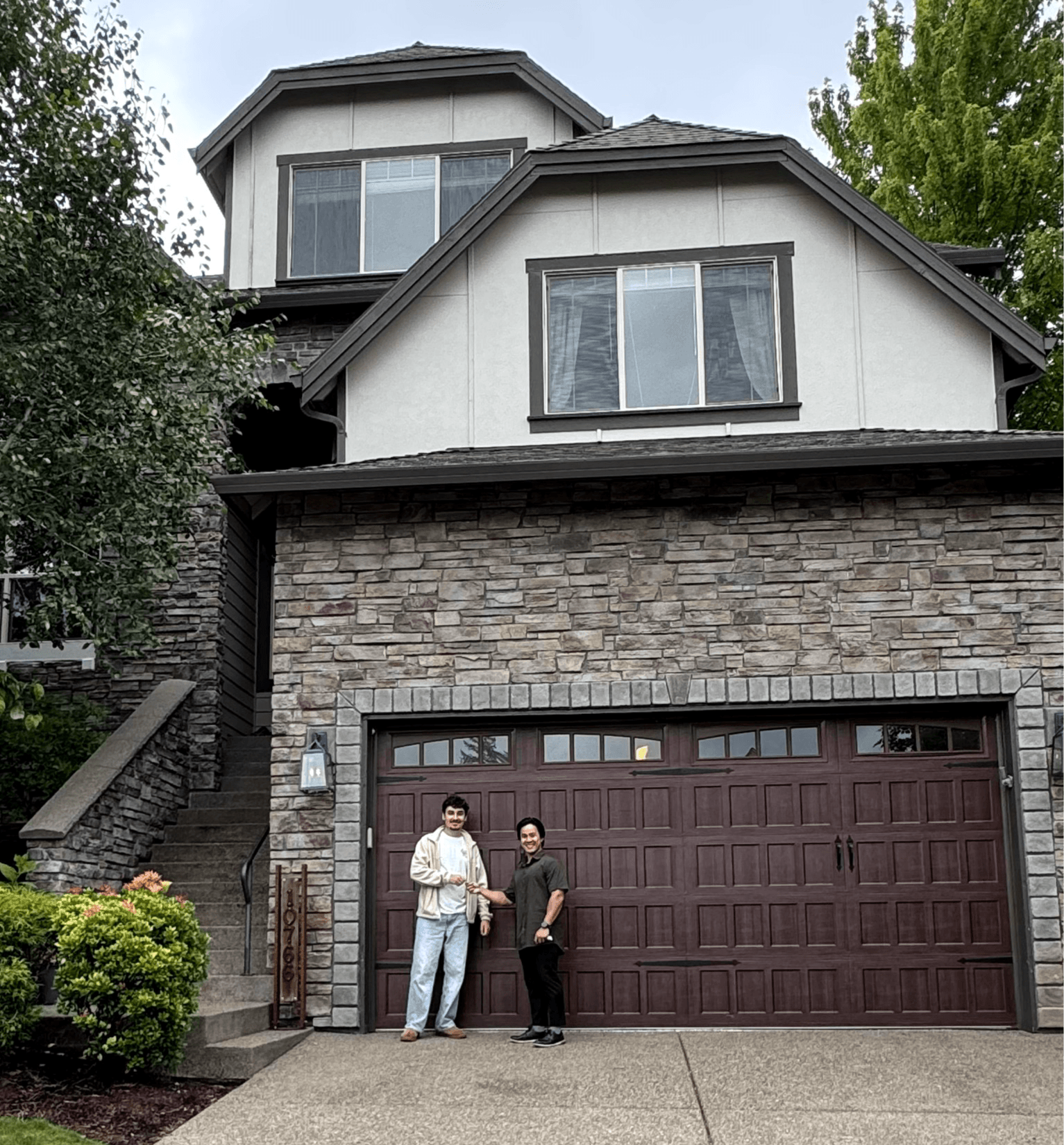 Two people standing in front of a house with a brick garage door, trees, and stairs visible, daytime.