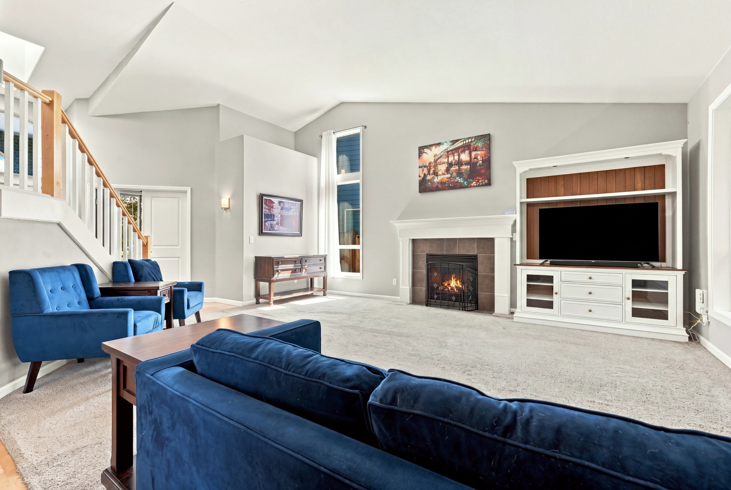 Living room with blue sofas, fireplace, TV, and artwork, bright with white walls and large windows.