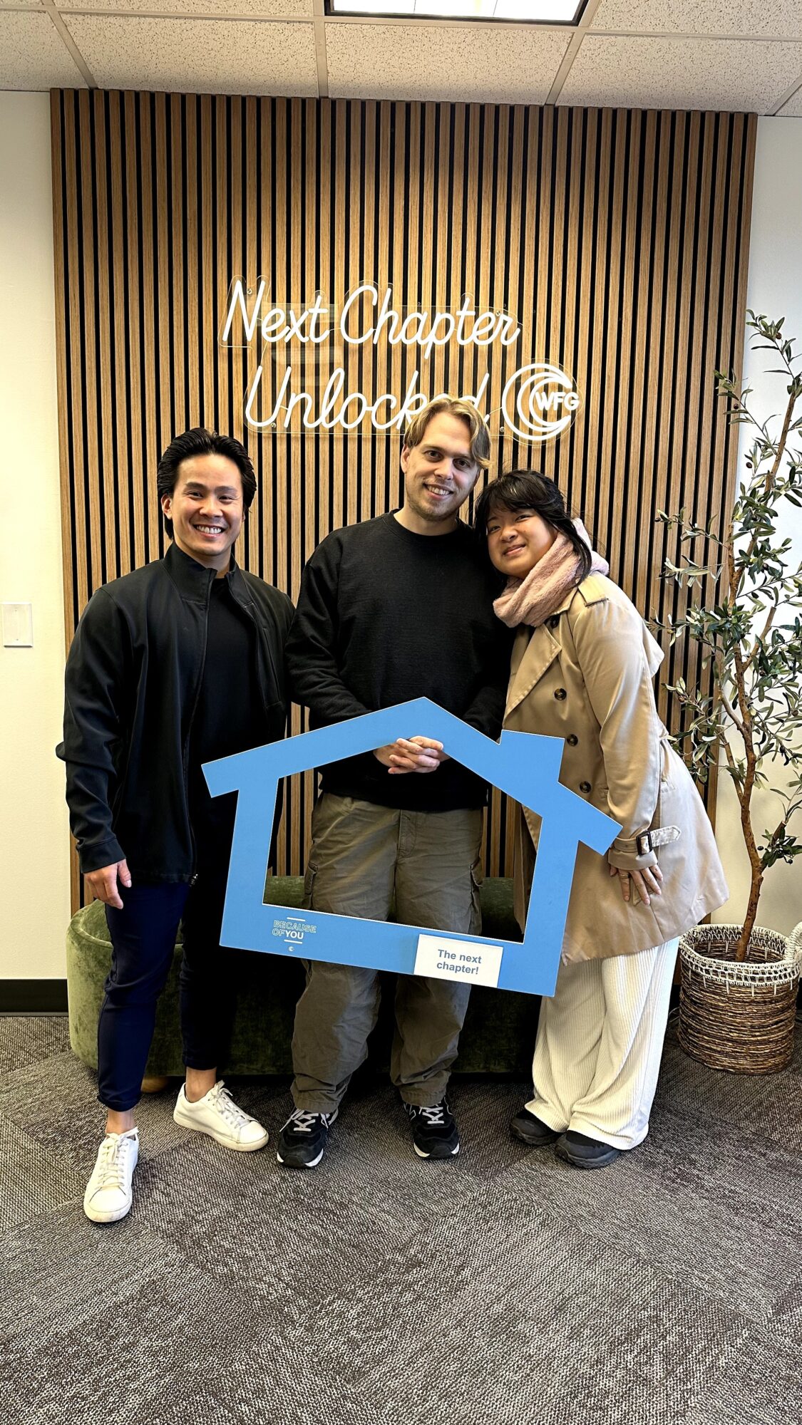 Three people standing indoors, holding a blue house-shaped frame, smiling, with a wooden wall and potted plant behind them.