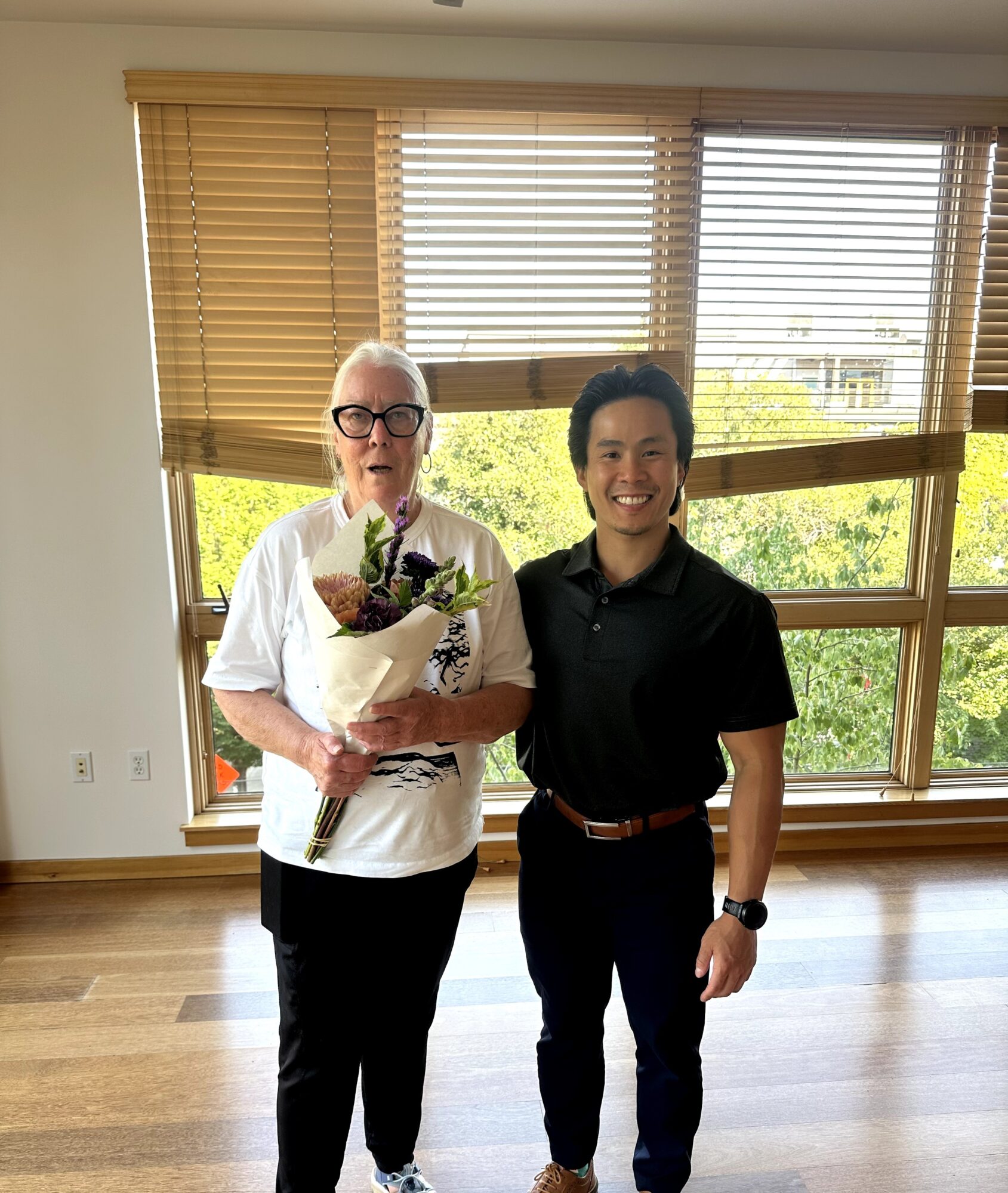 Two people standing indoors in front of large windows with blinds, smiling, one holding a bouquet of flowers.