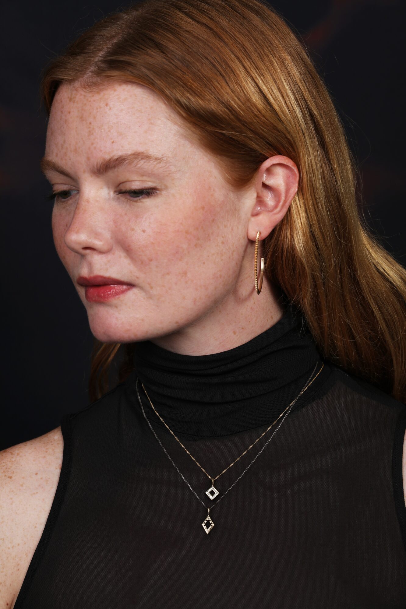 Side profile of a woman with red hair, wearing hoop earrings and layered necklaces, against a dark background.
