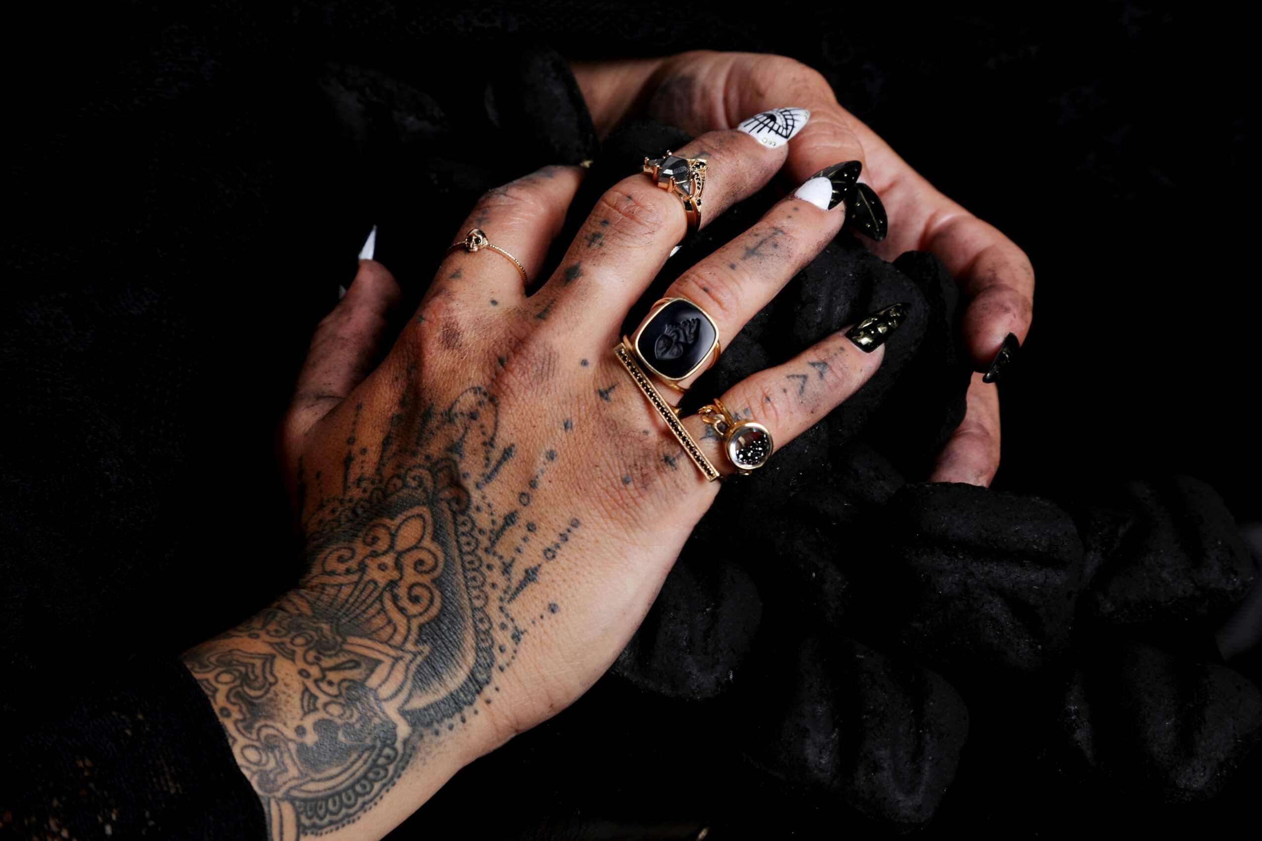 Close-up of hands with tattoos and rings, holding black fabric against a dark background.