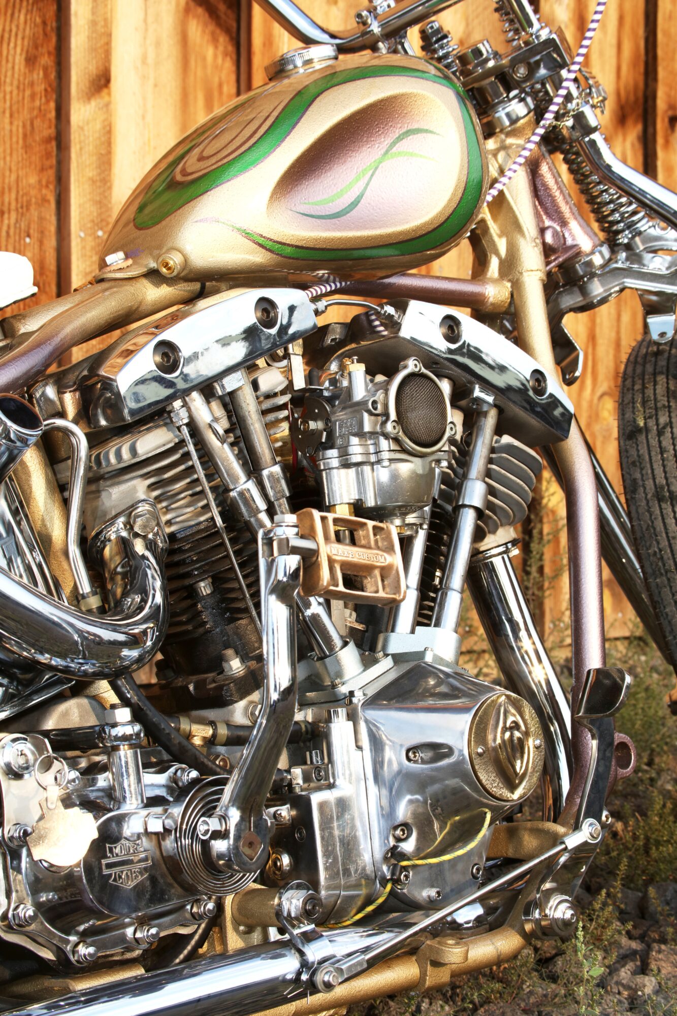 Close-up of a motorcycle engine and fuel tank with green and beige paint, wooden background, and chrome details.