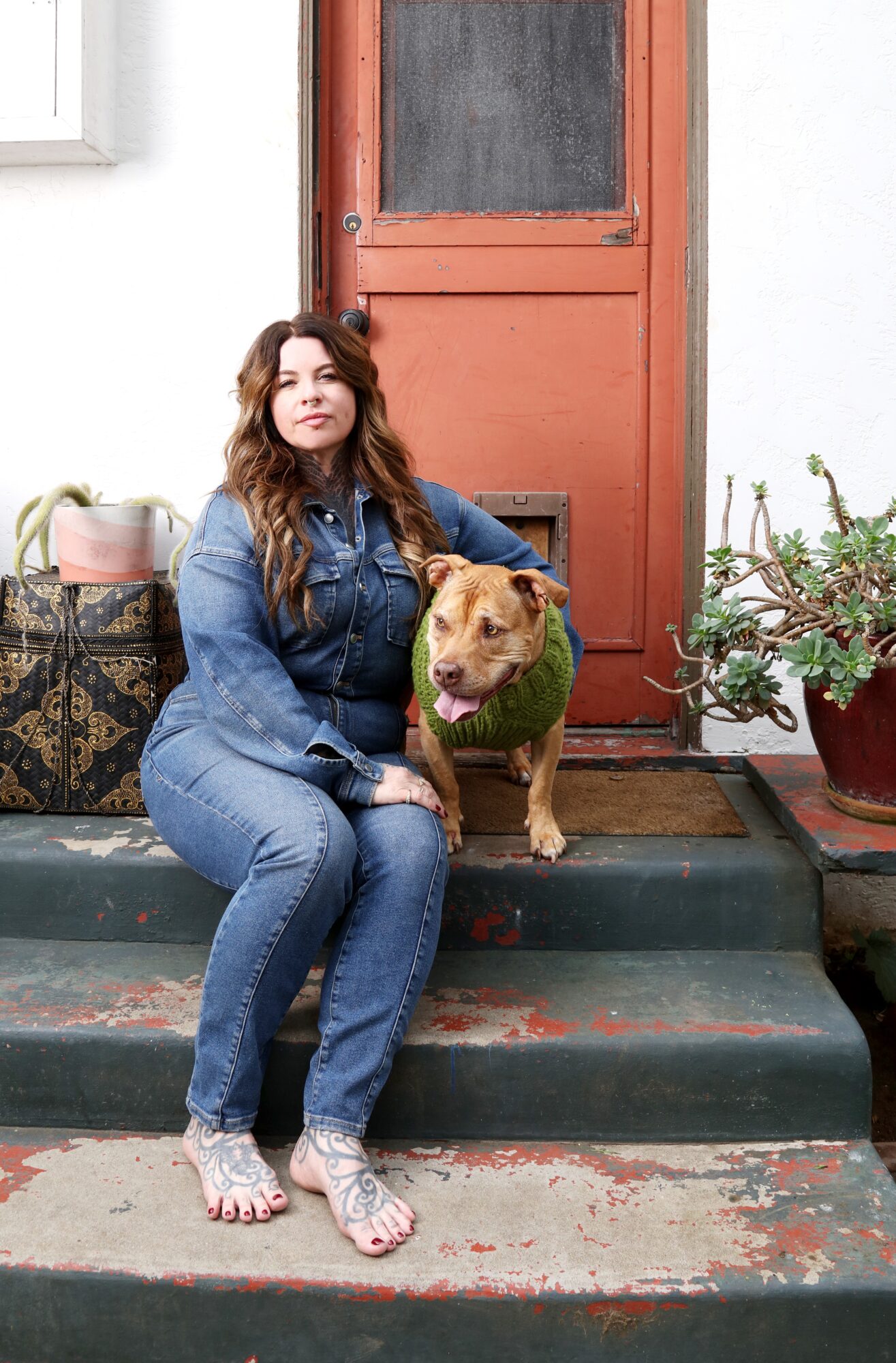 Woman sitting on steps with dog wearing a green sweater, door and potted plants in background.