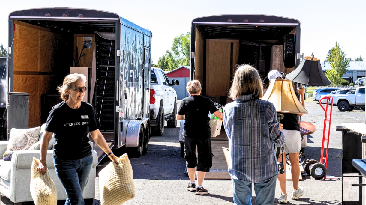 People unload items from two trucks in a parking lot, with trees and vehicles in the background.