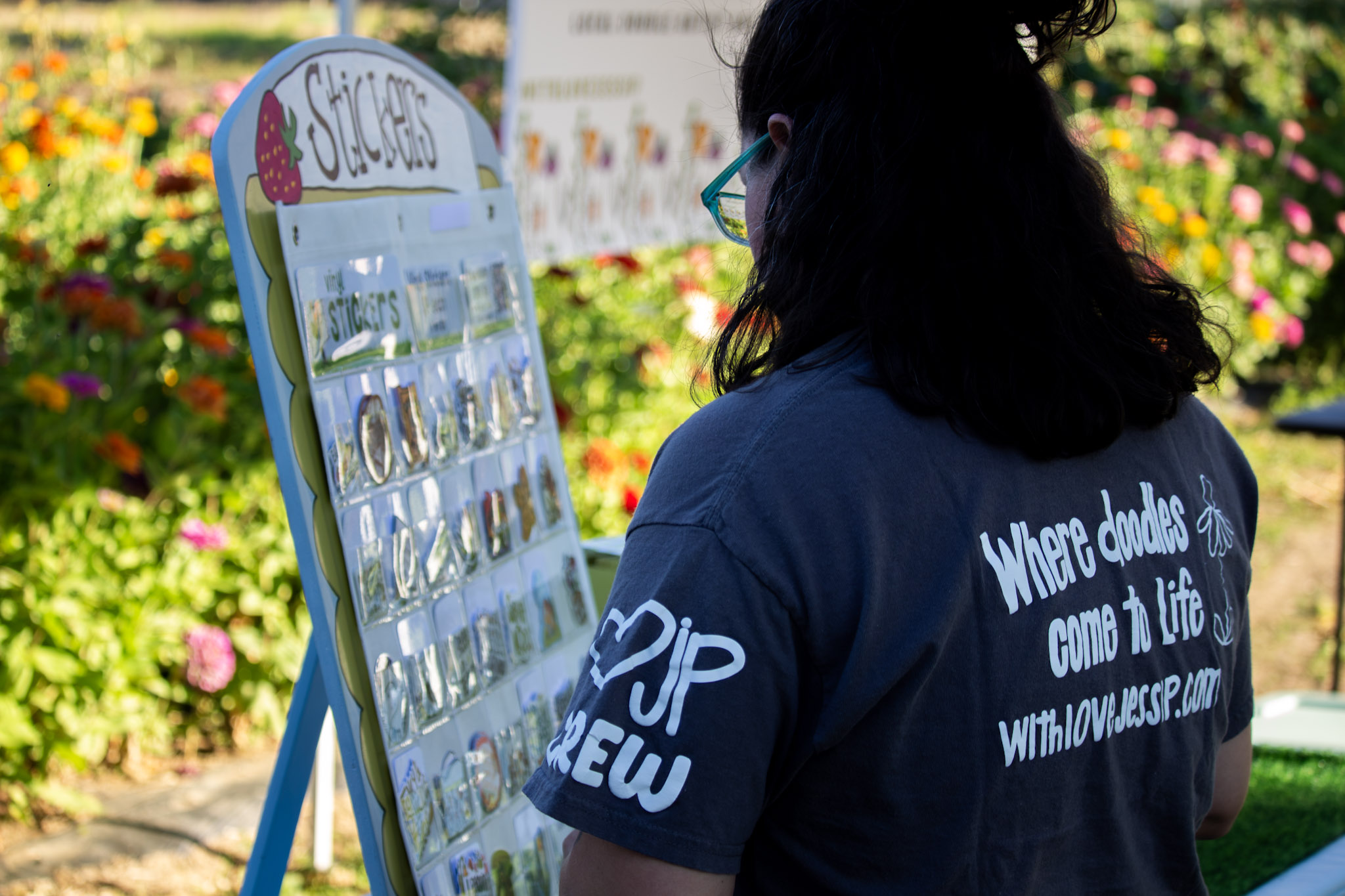 Person with dark hair and glasses looking at a display board outdoors, surrounded by colorful flowers.
