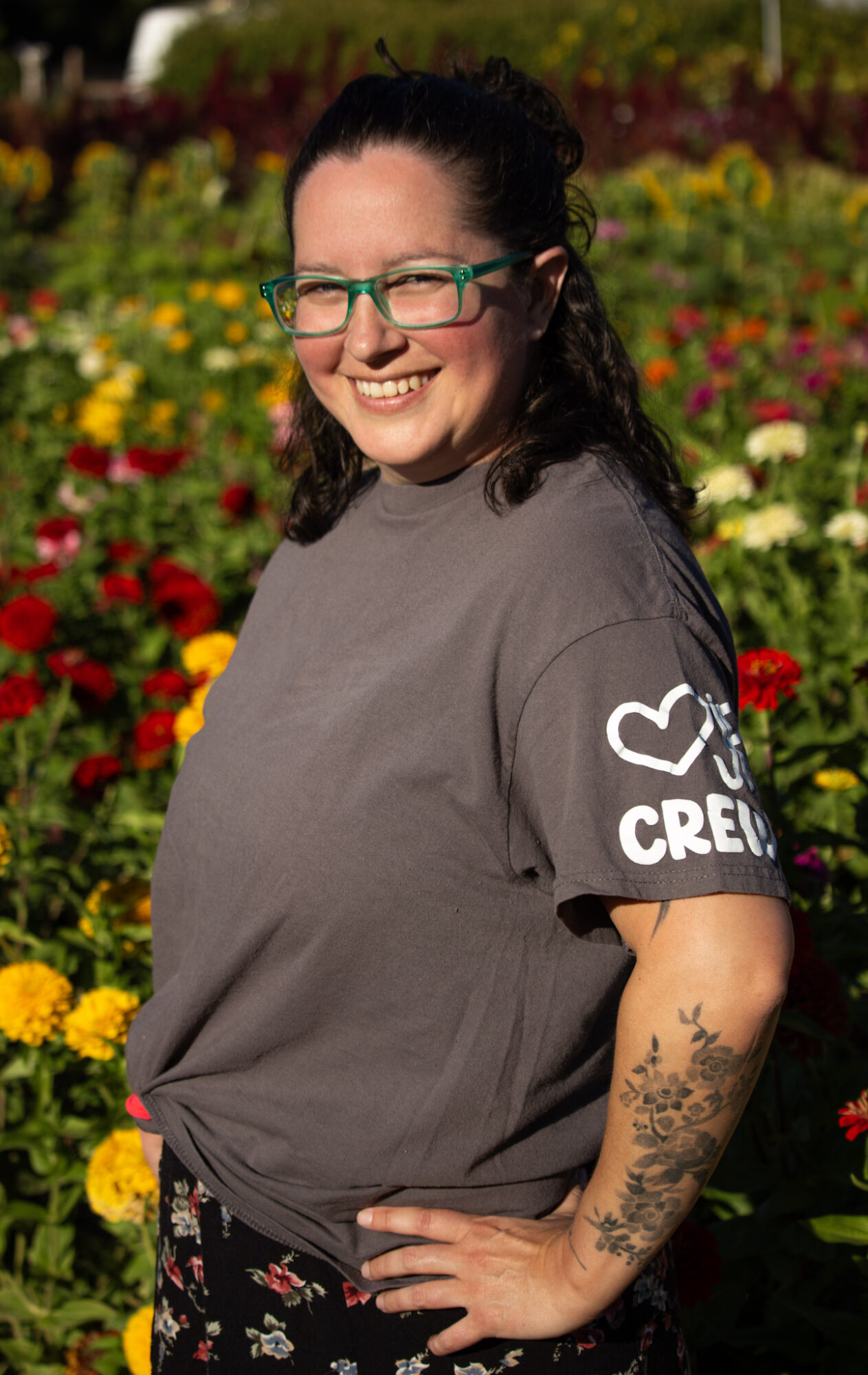 Young woman with glasses smiling outdoors in a colorful flower garden, wearing a gray T-shirt with a heart and text on sleeve.
