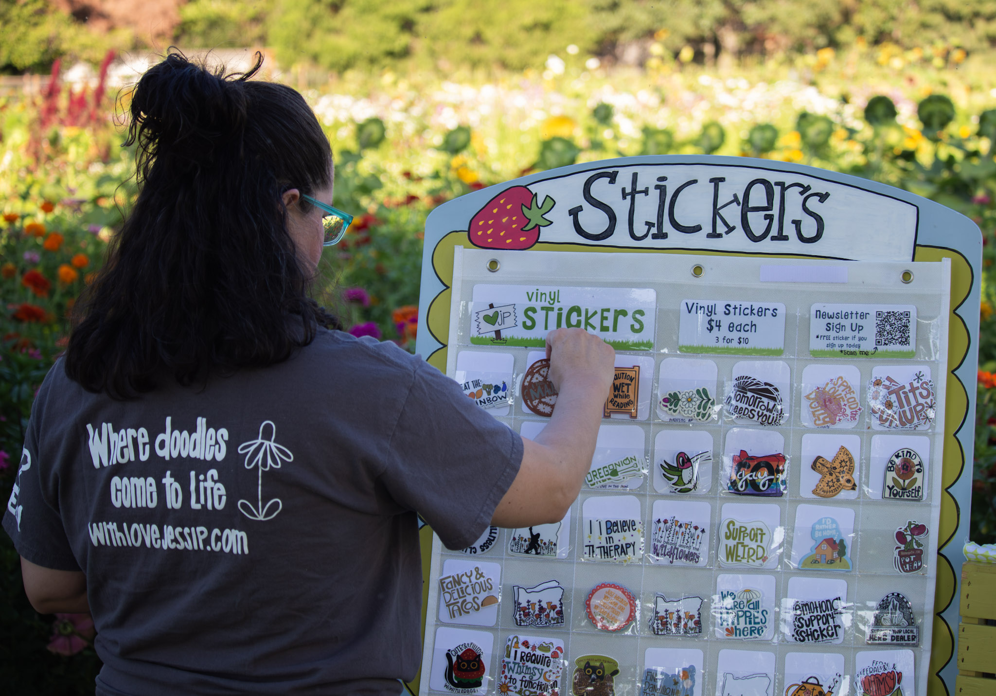 Person with dark hair and glasses selecting stickers from a display board outdoors, with colorful flowers in the background.