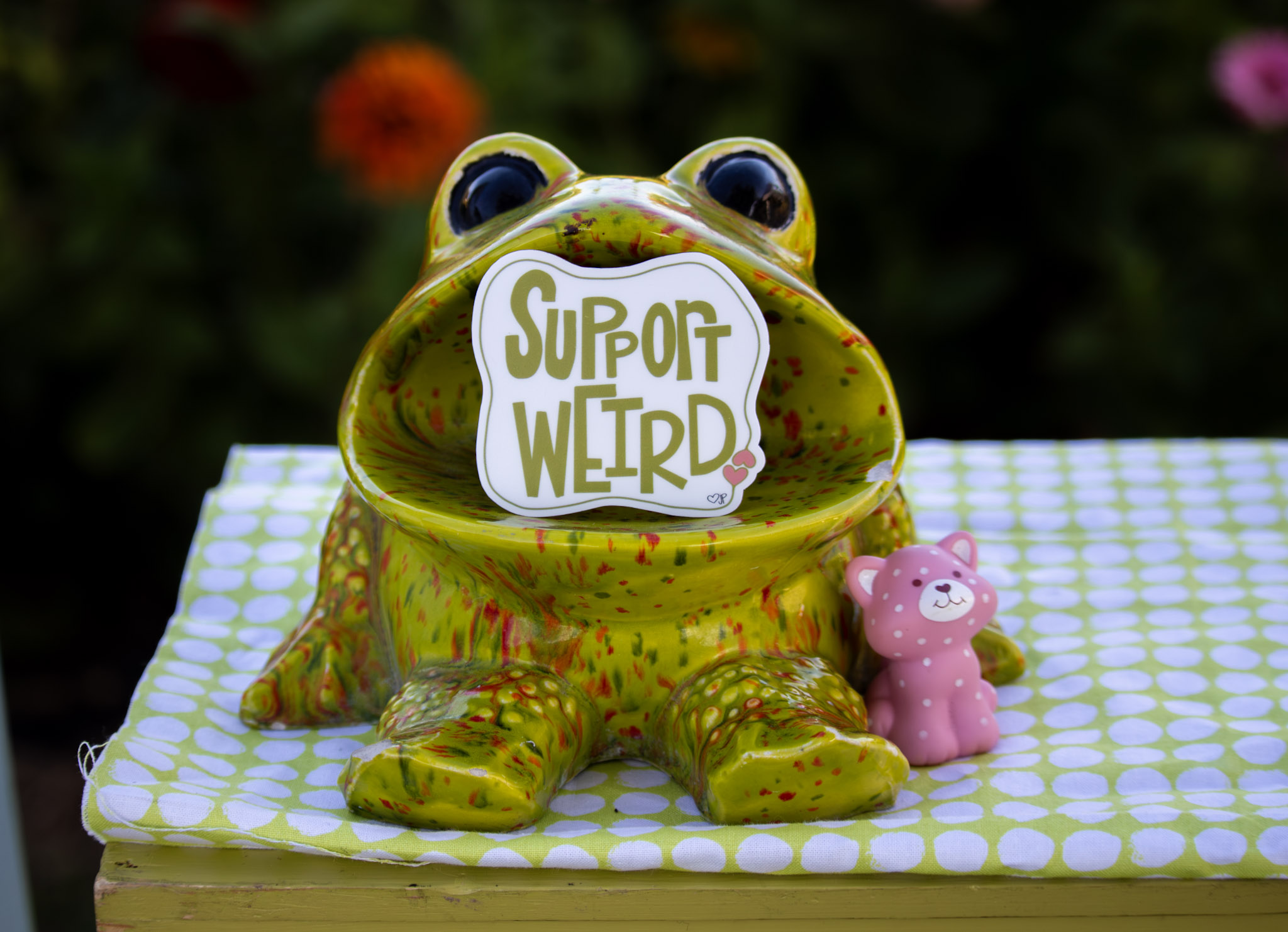 Green frog-shaped figurine with a sign reading 'Support Weirdo' in its mouth, sitting on a checkered surface with a small pink pig figure nearby, outdoors.