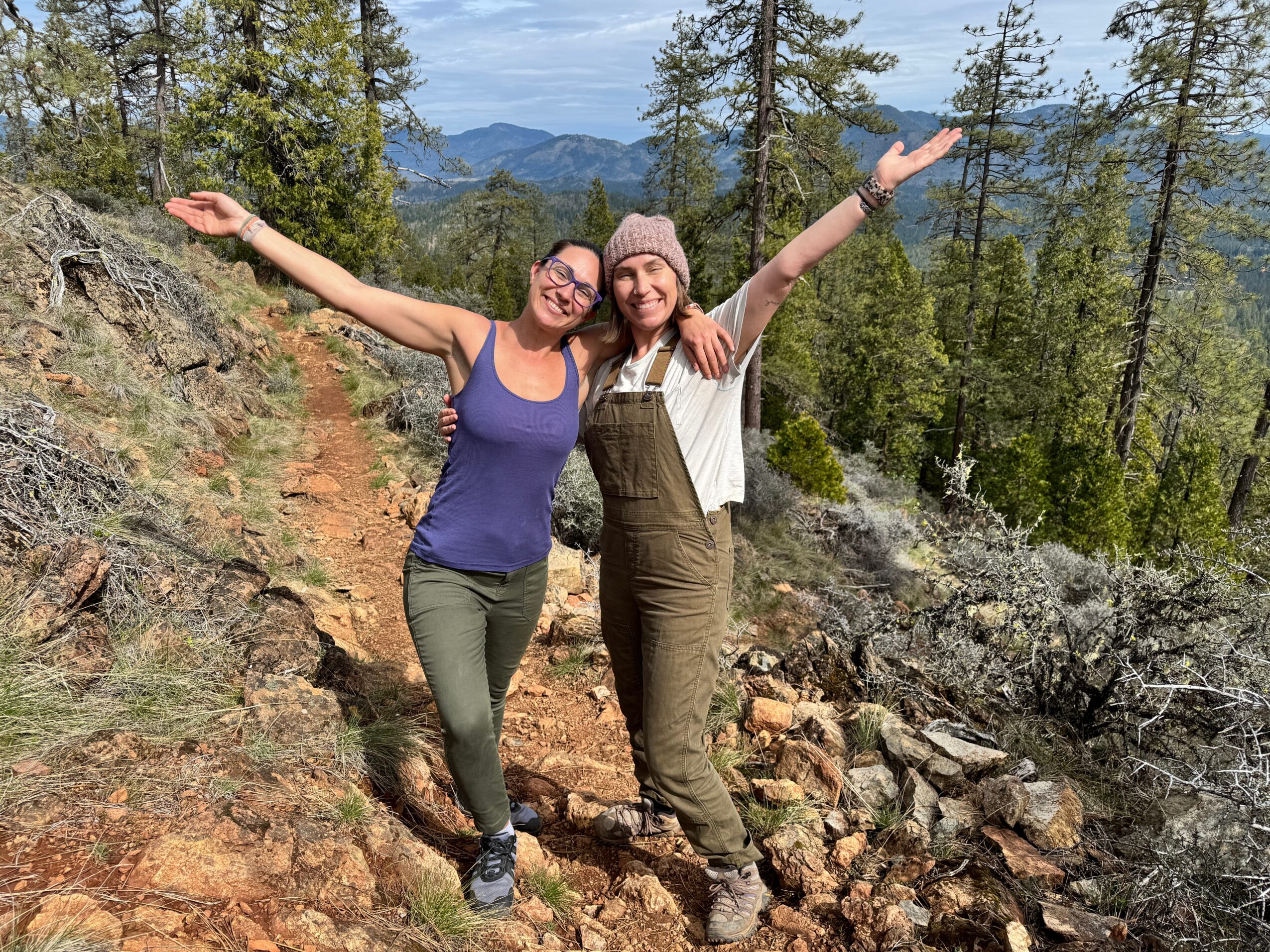 Two women stand on a rocky trail in a forested mountain area, smiling with arms raised, surrounded by trees and mountains.