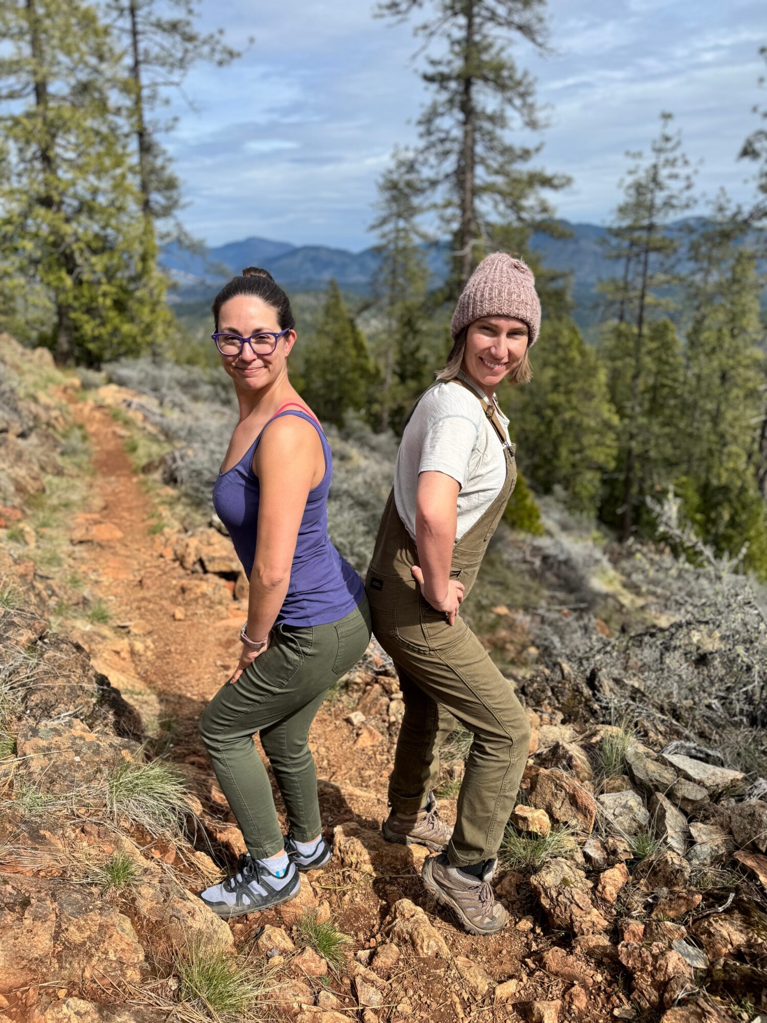 Two women hiking on a rocky trail in a forested area with mountains in the background, smiling at the camera.