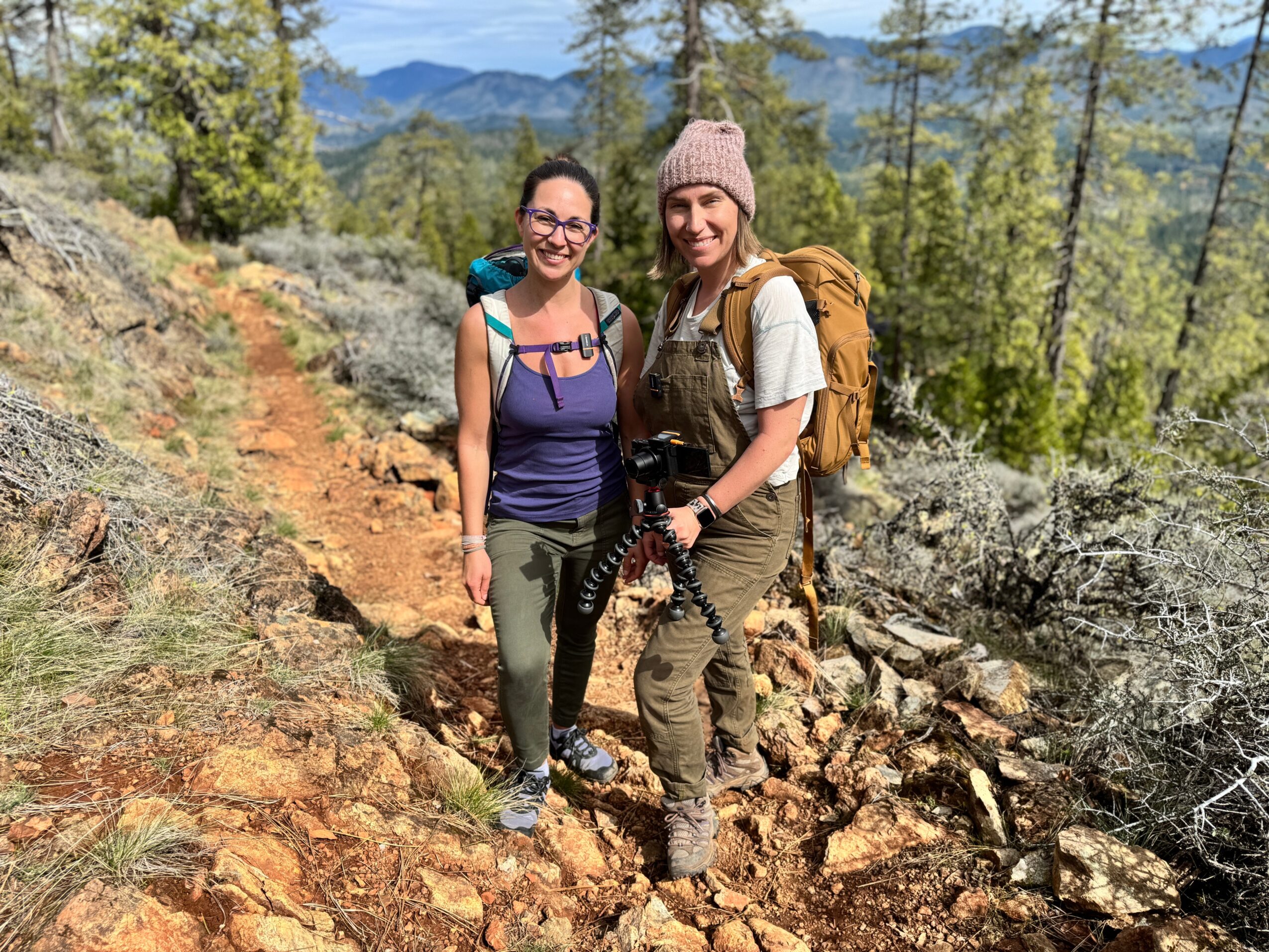 Two women hiking on a rocky trail in a forested mountainous area, smiling, with trees and mountains in the background.