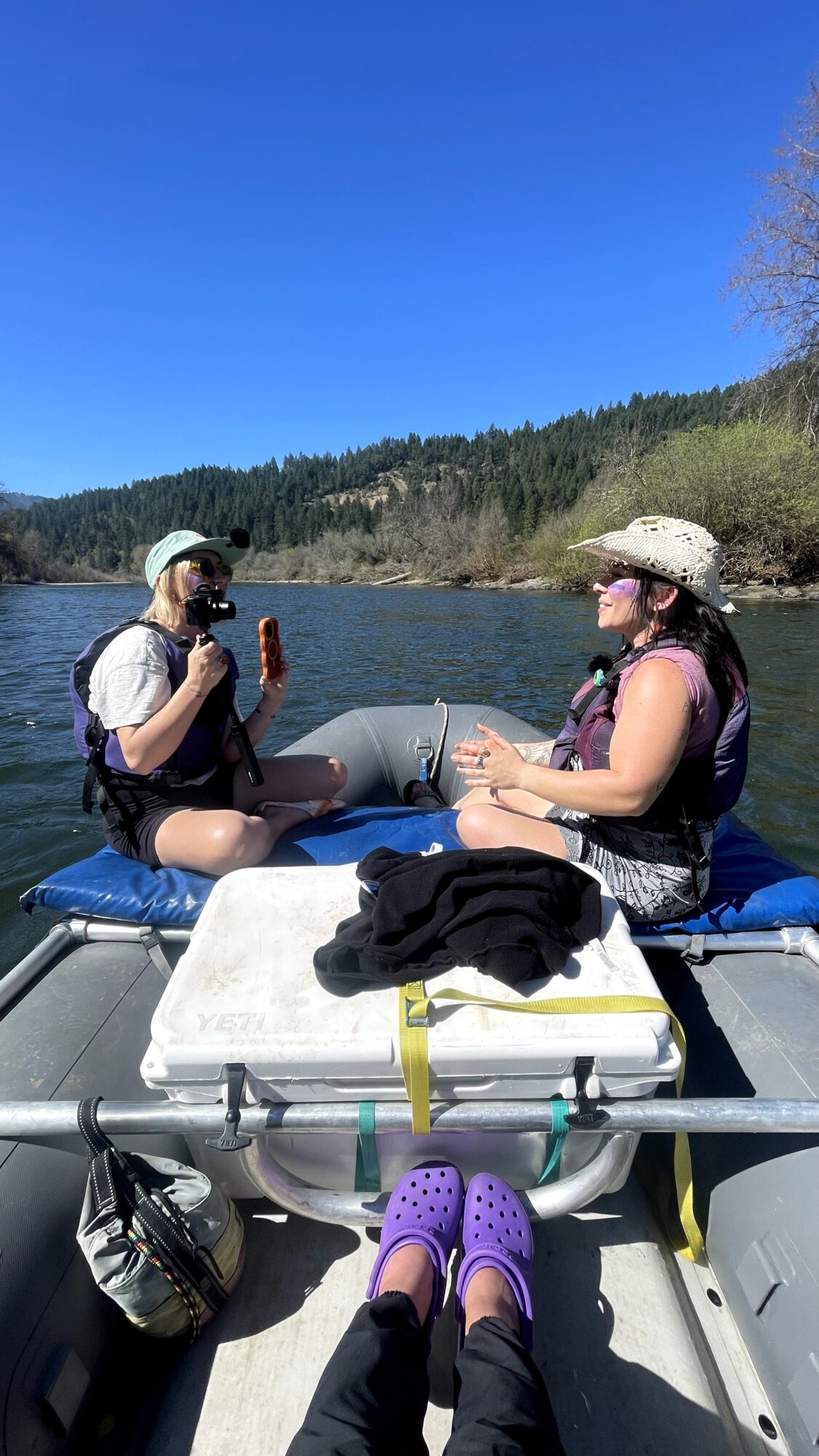 Two women sit in a boat on a river, wearing hats and sunglasses, with a forested landscape in the background.