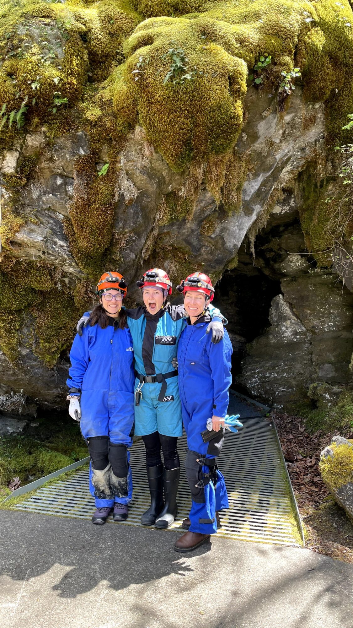 Three people in blue jackets and hats standing on a metal platform in front of a moss-covered rock formation.