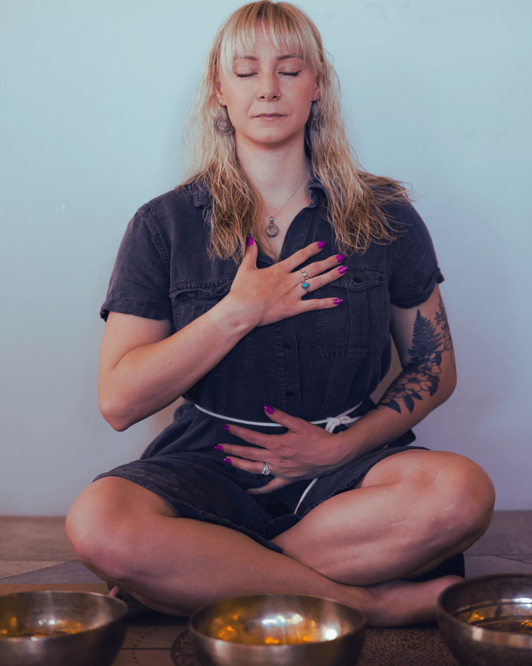 Woman sitting cross-legged with eyes closed, hand on chest, and other hand on stomach, against a plain background.