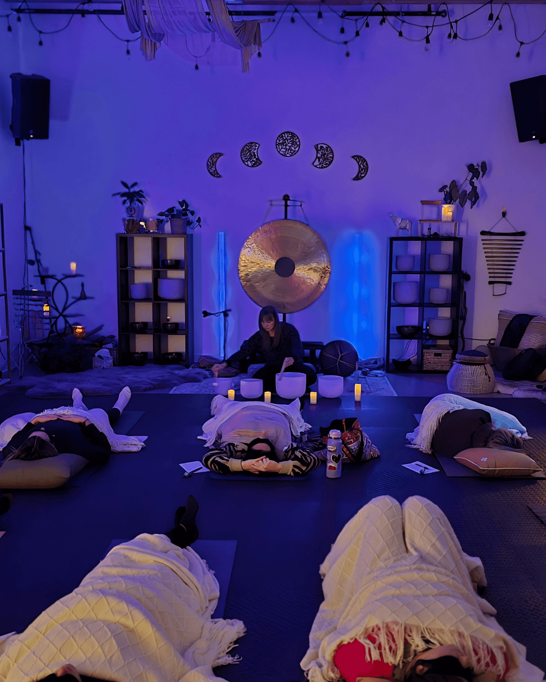 People lying on mats with candles in a dimly lit room decorated with plants, shelves, and moon phase symbols, with a person sitting in the center.