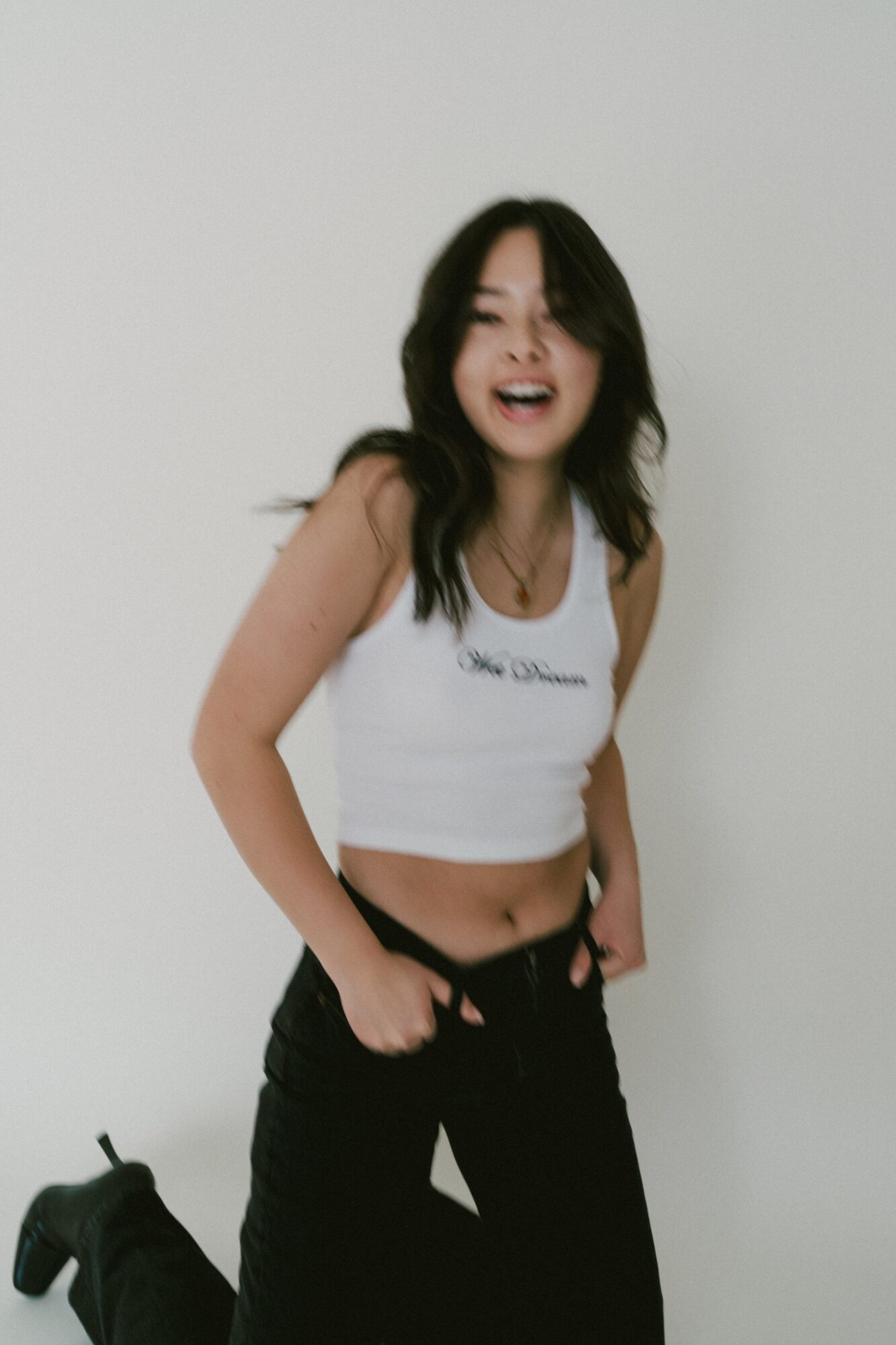Young woman with long dark hair, smiling and laughing, wearing a white crop top and black pants, standing against a plain white background.