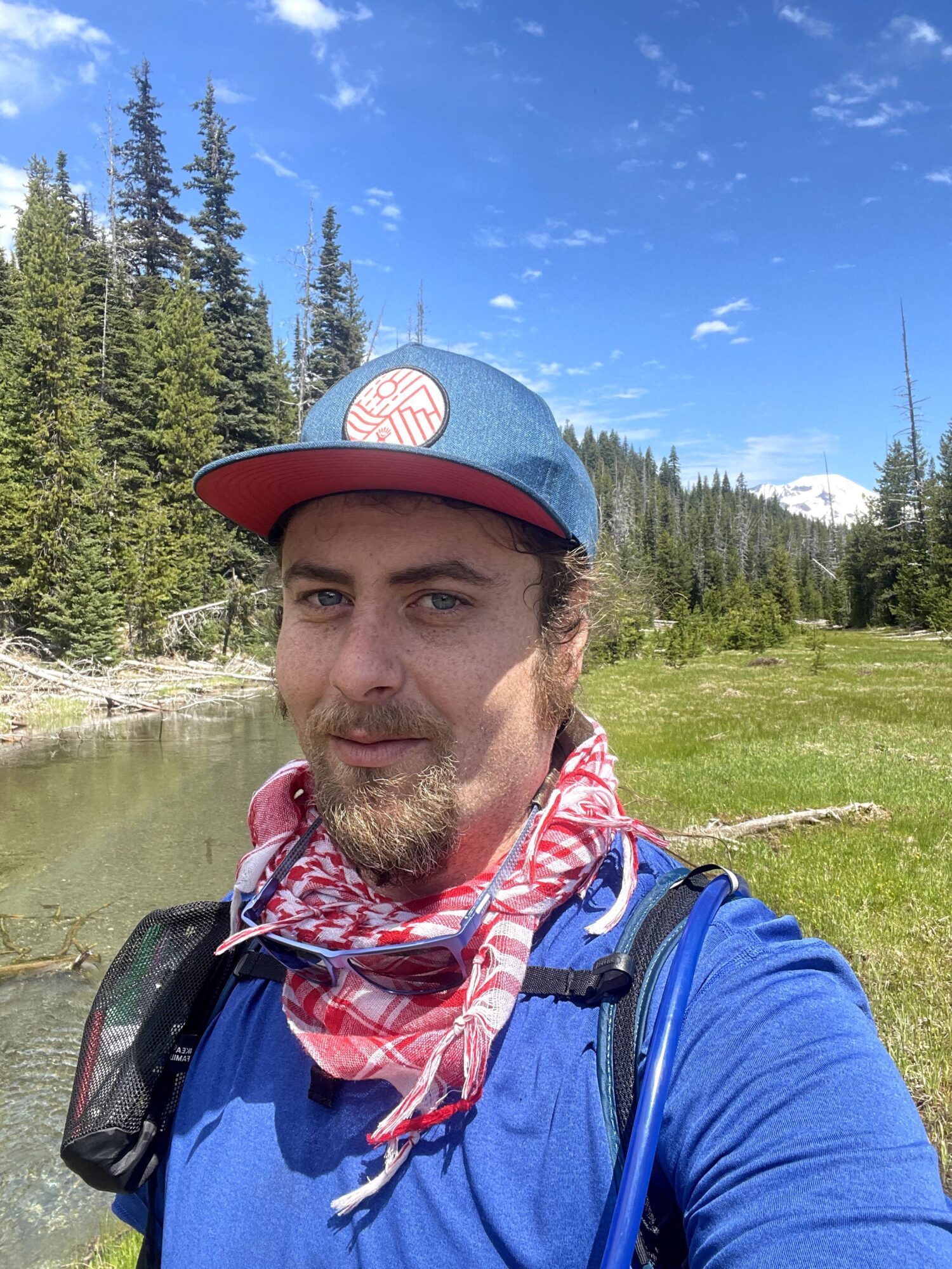 Man outdoors in a blue shirt and cap, with a red and white scarf, standing near a river with trees and blue sky.