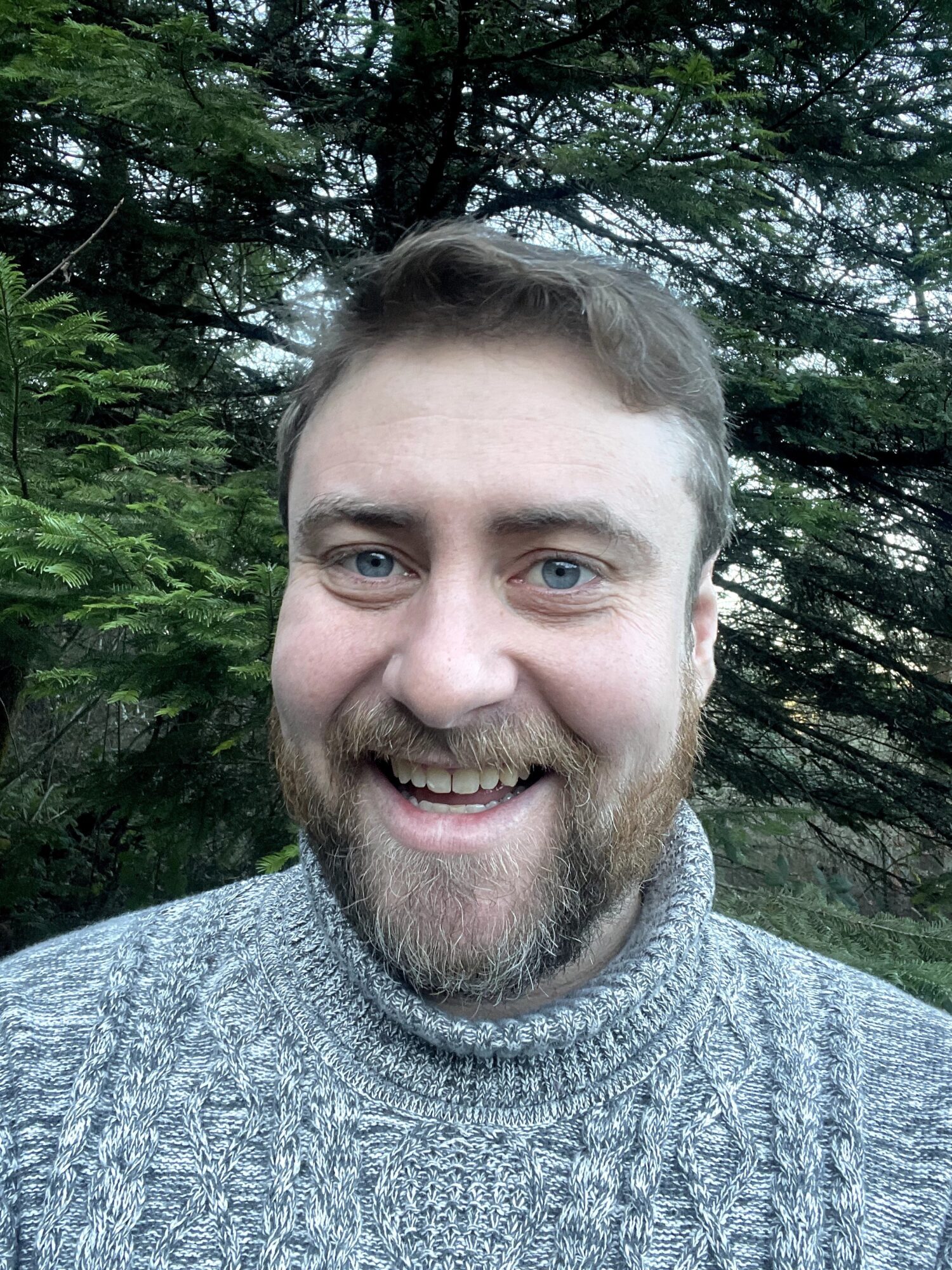 Man with a beard smiling outdoors, surrounded by trees, wearing a gray sweater.