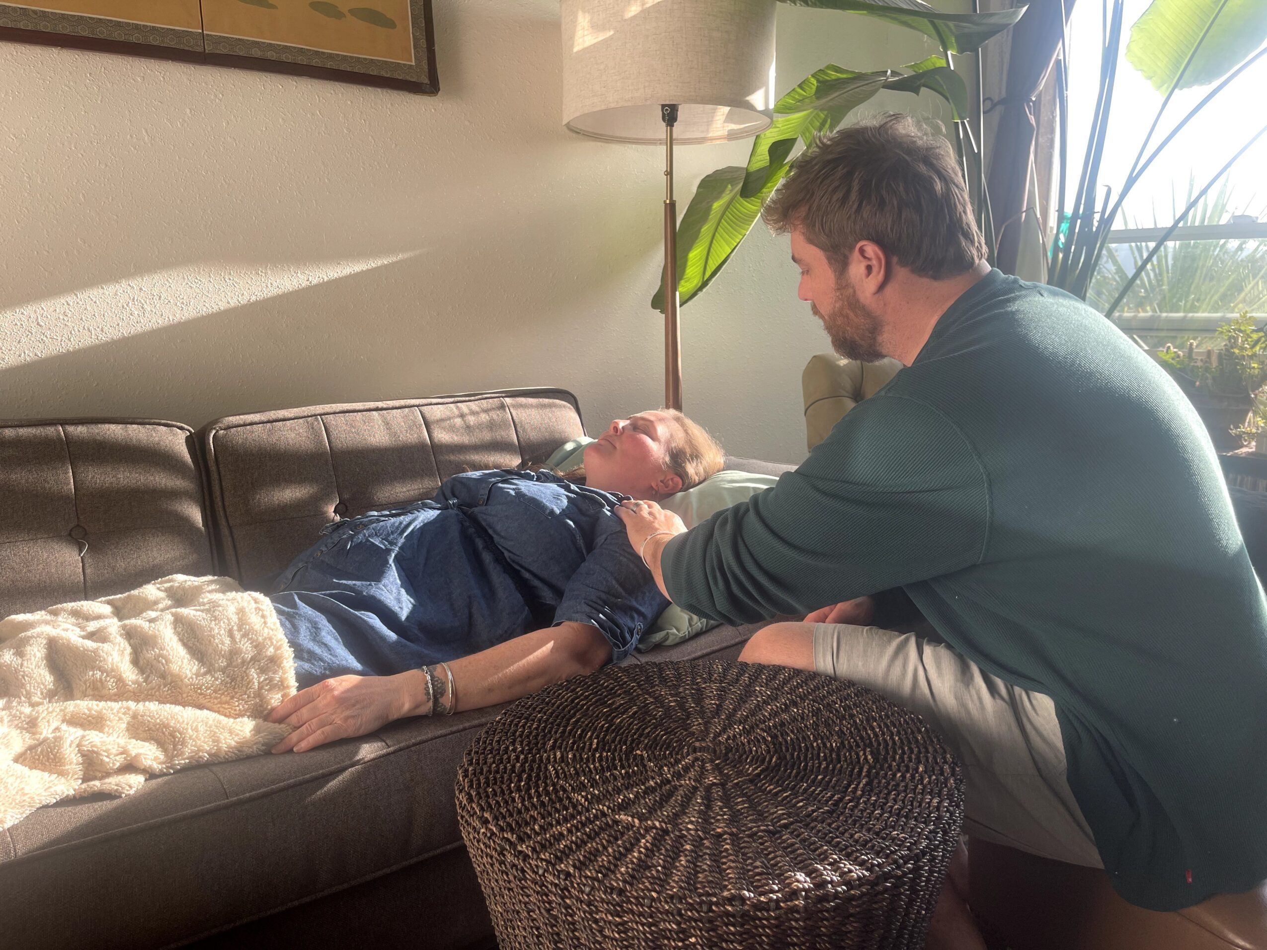 A man sitting beside a woman lying on a sofa, holding her hand and looking at her, in a living room.