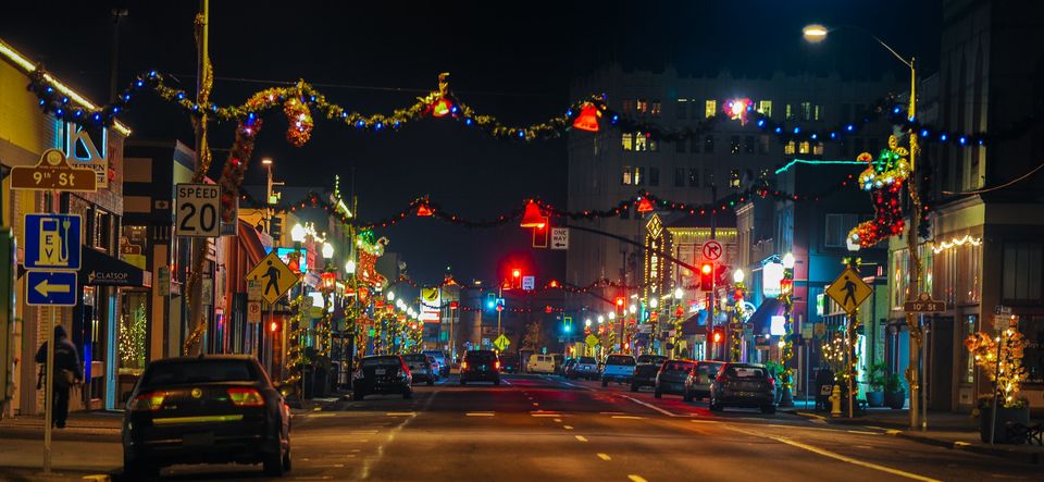 City street at night decorated with colorful holiday lights, with cars parked along the sides and buildings illuminated.
