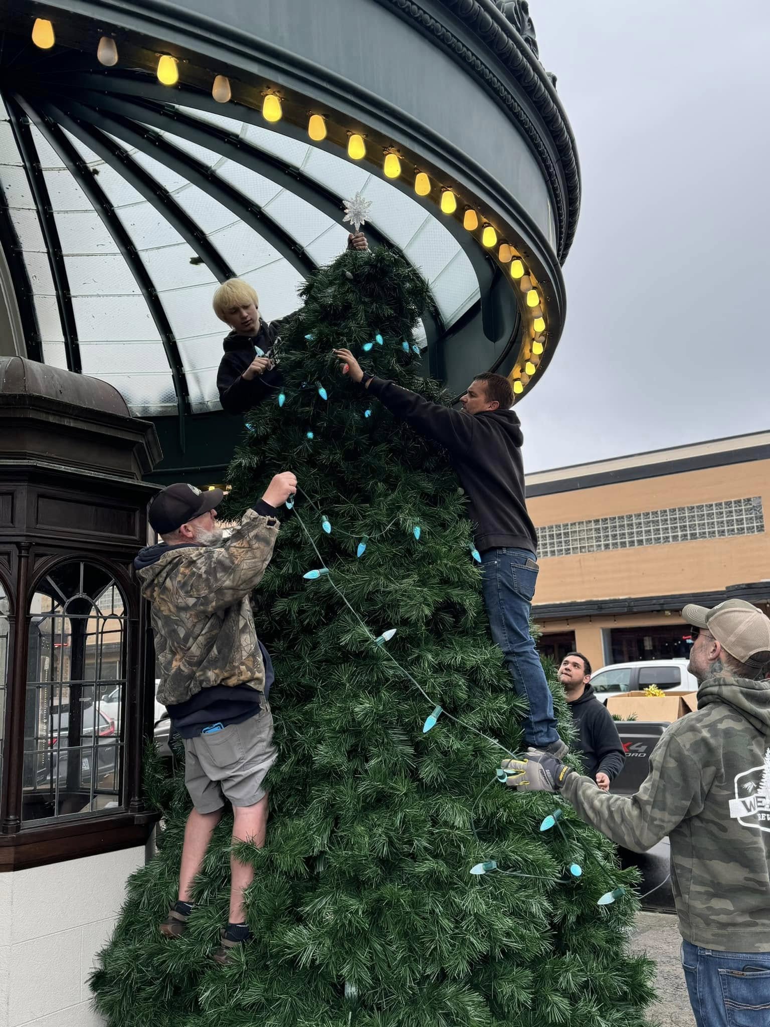 People decorate a large Christmas tree outside a building with a glass dome, hanging ornaments and lights, during daytime.