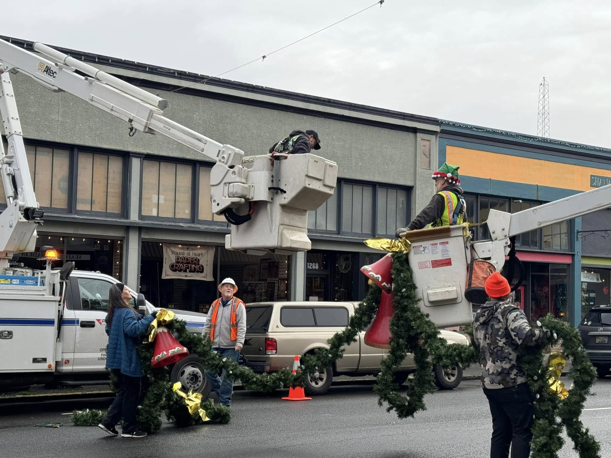 Workers decorate a street with Christmas garlands and wreaths, using a cherry picker to reach high areas, in front of a building.