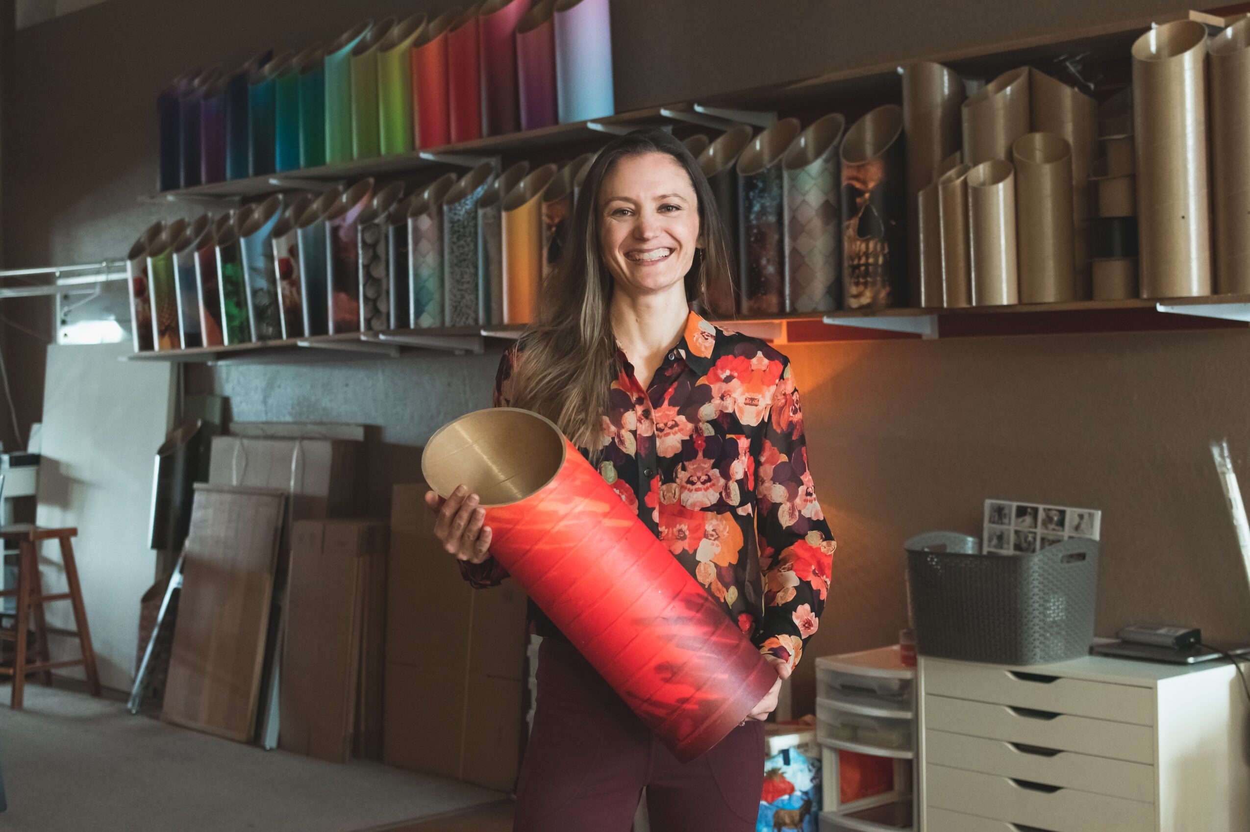 Woman holding a large red and gold cylindrical object, smiling, in a store with colorful rolls of material on shelves behind her.
