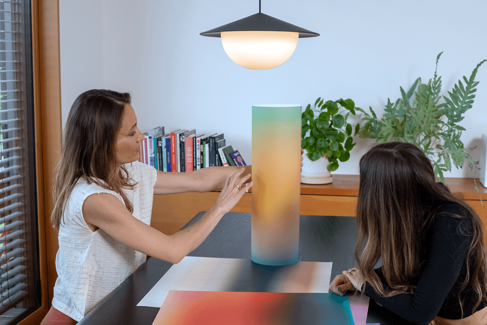 Two women sit at a table with a colorful lamp, books, and plants in a bright room.