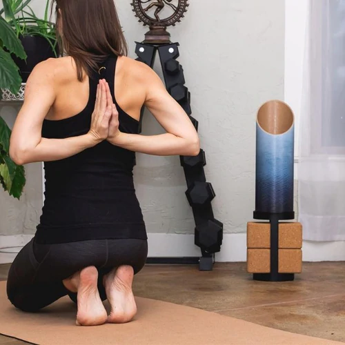 Person kneeling on yoga mat with hands in prayer position, practicing yoga indoors near decorative objects.