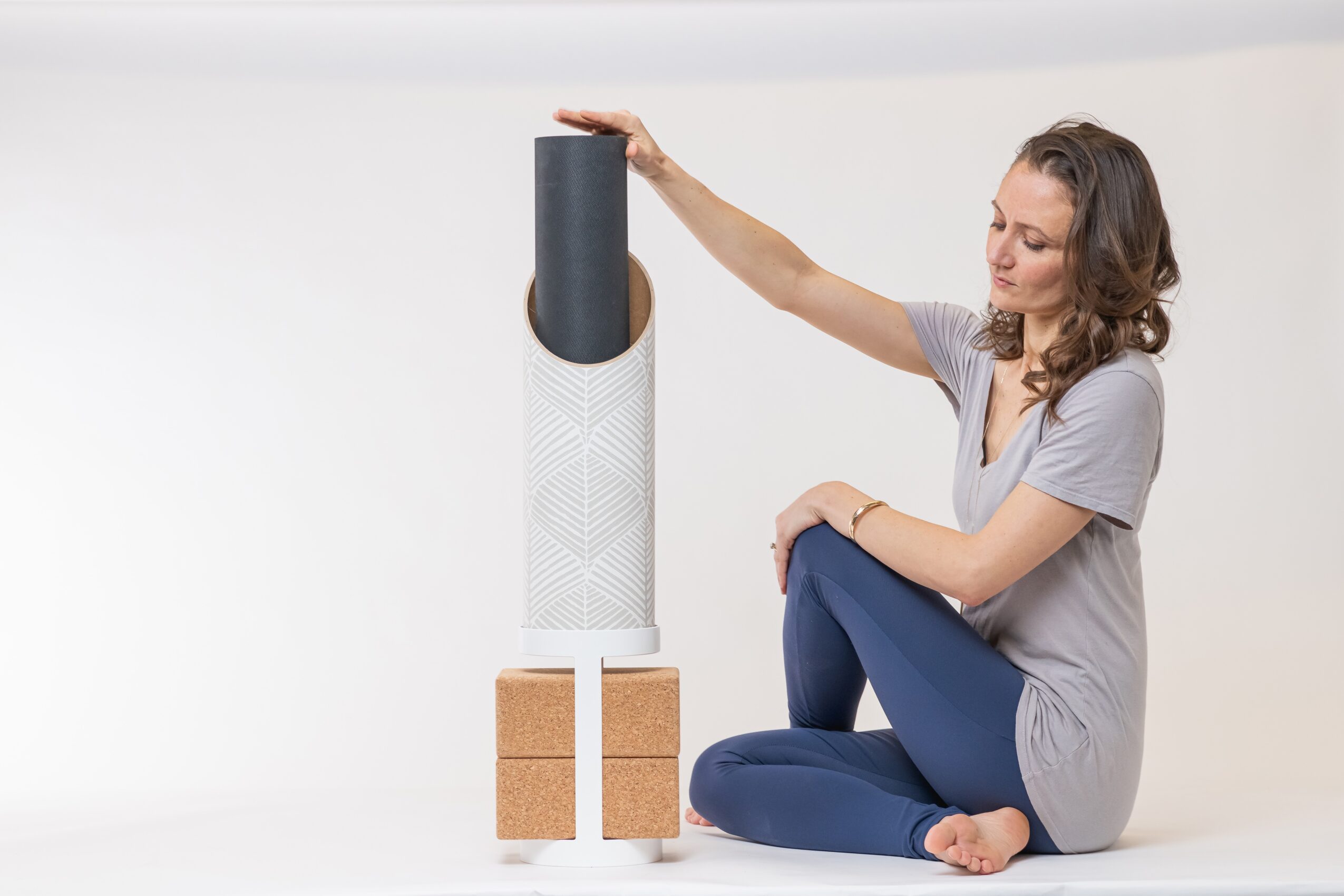 Woman sitting cross-legged on the floor, reaching out to a tall air purifier with a black top, white body, and cork blocks at the base.