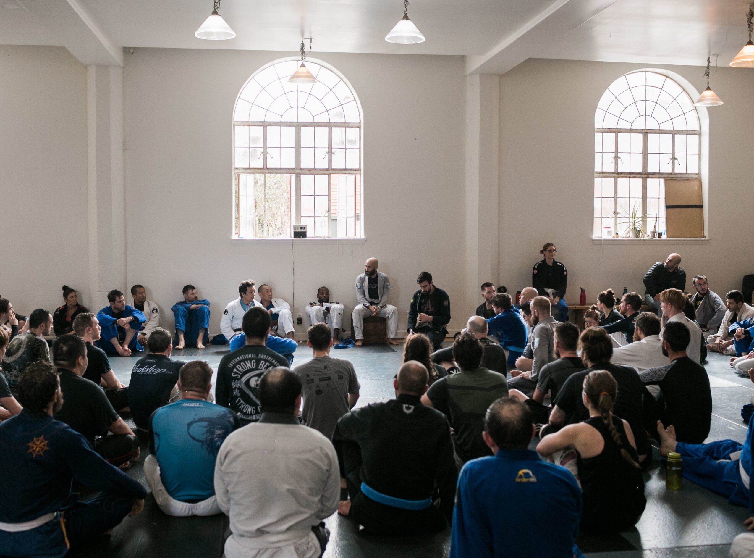 A large group sits in a circle inside a bright room with arched windows.