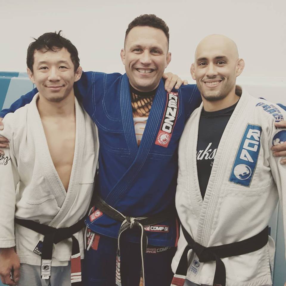 Three people wearing martial arts uniforms stand together with their arms around each other.