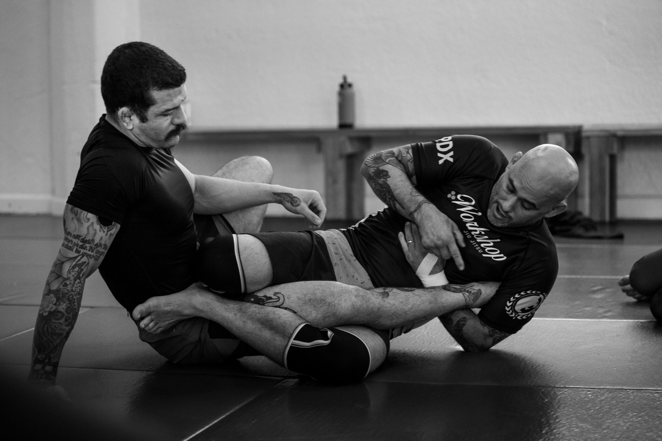 Two people practice grappling on a mat in a gym.
