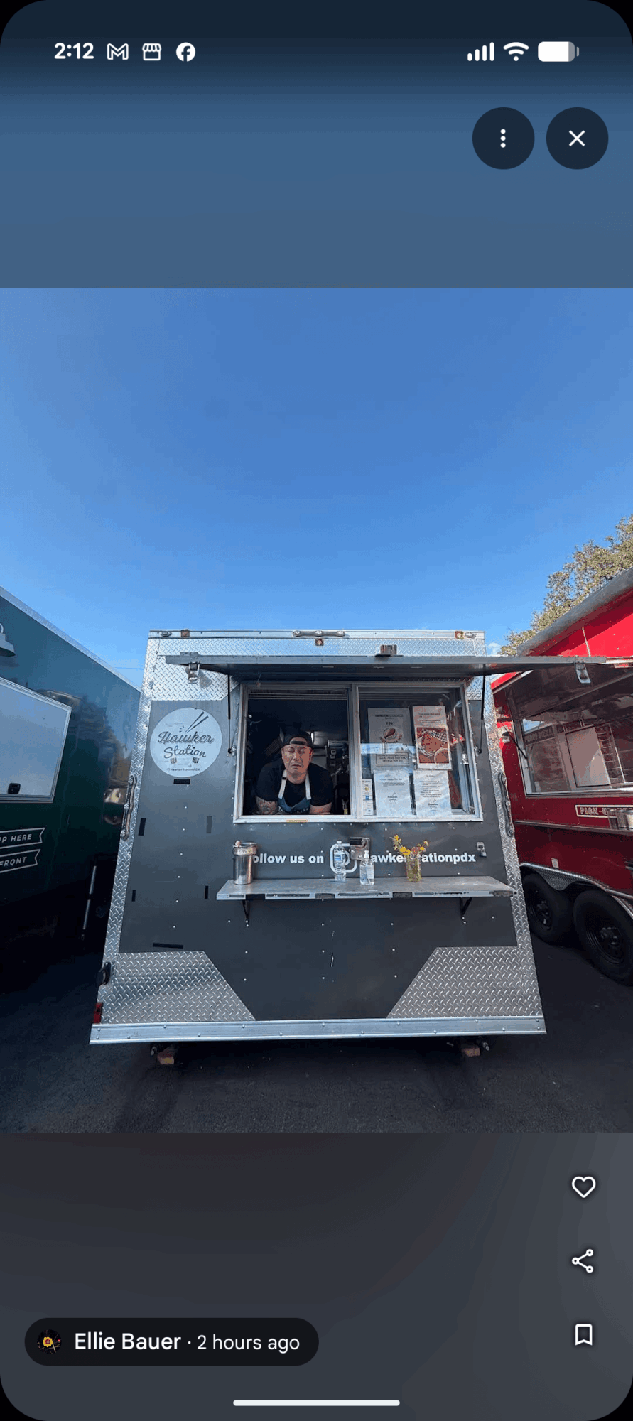 A silver food truck with an open service window is parked between two other trucks beneath a blue sky.