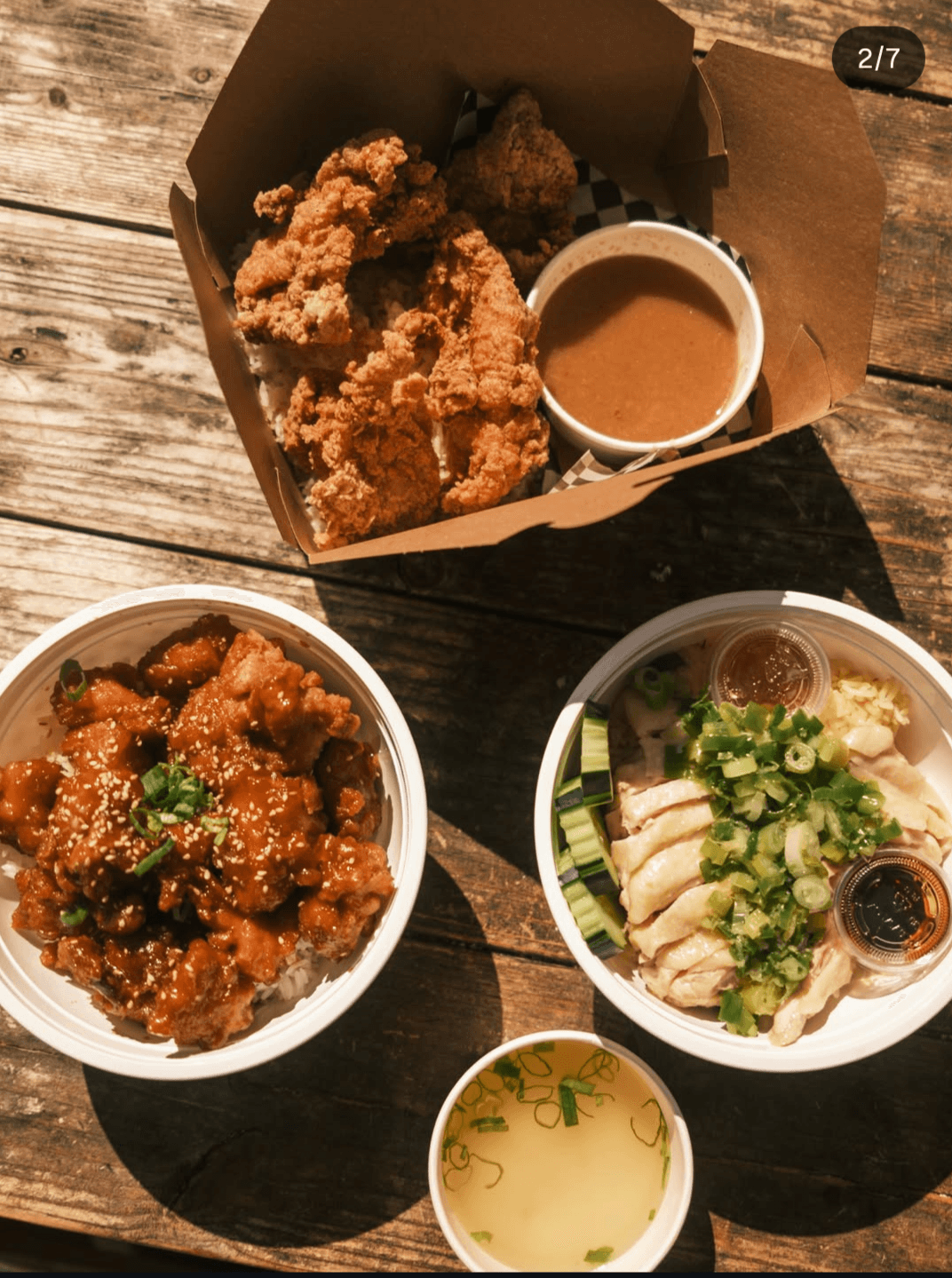 Takeout meal with fried chicken, two noodle dishes, dipping sauces, and a cup of broth on a wooden table.