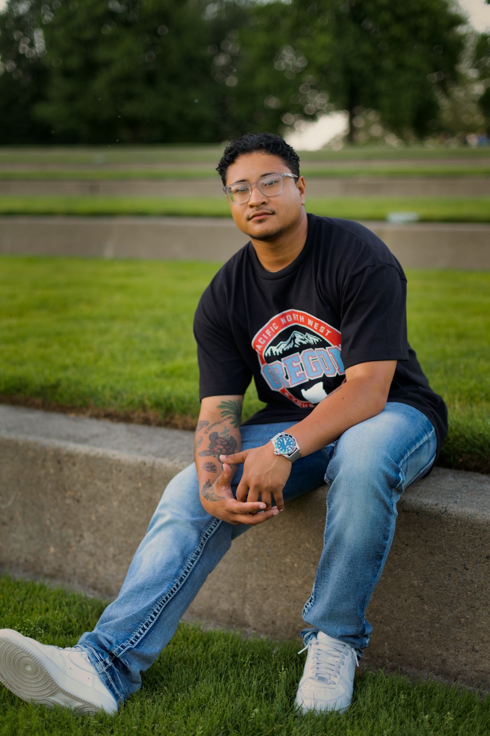 Young man sitting on a concrete ledge outdoors, wearing glasses, a black t-shirt, and jeans, with trees in the background.