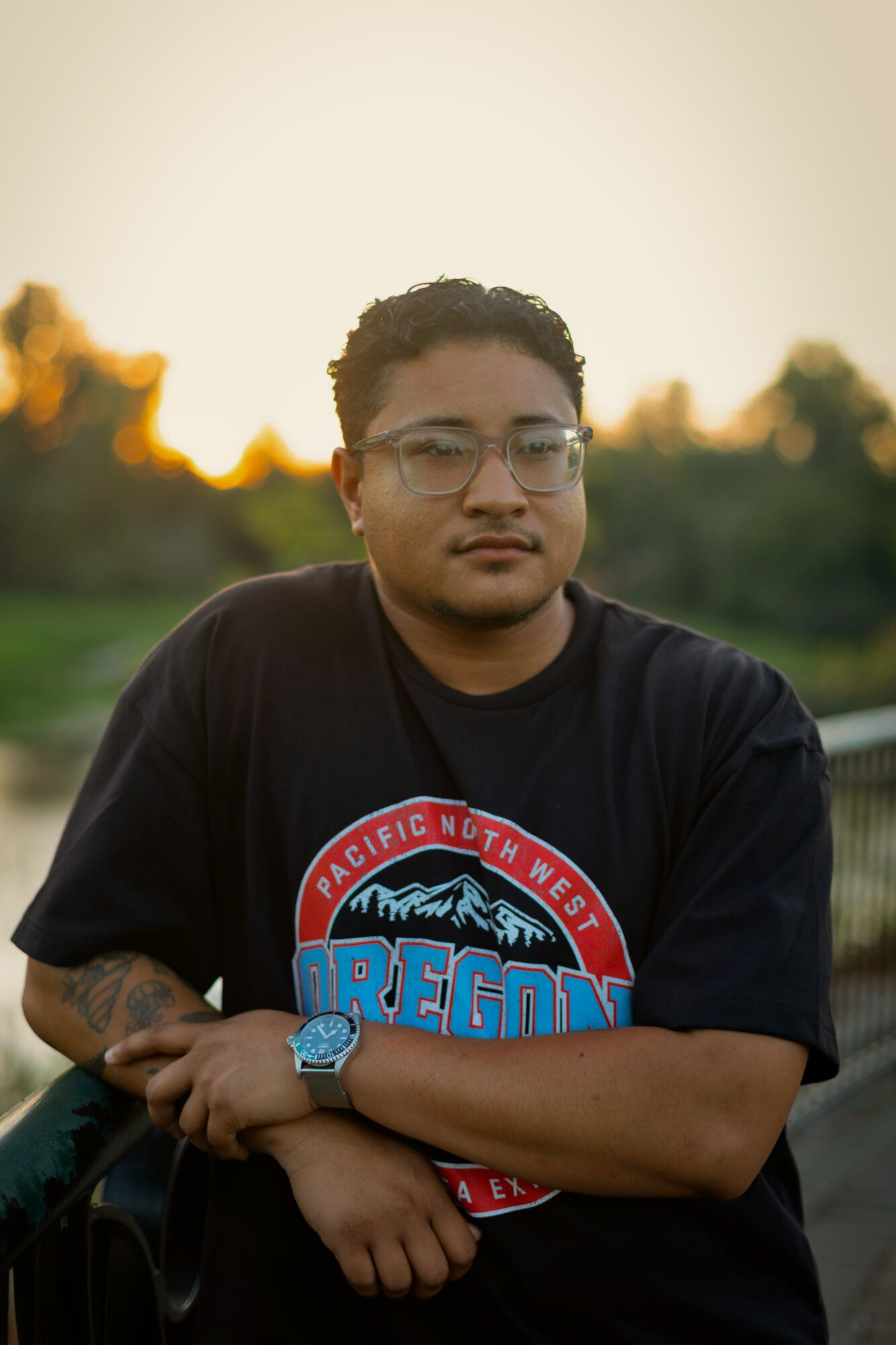 Young man with glasses and curly hair outdoors, wearing a black T-shirt with Oregon logo, standing with arms crossed.