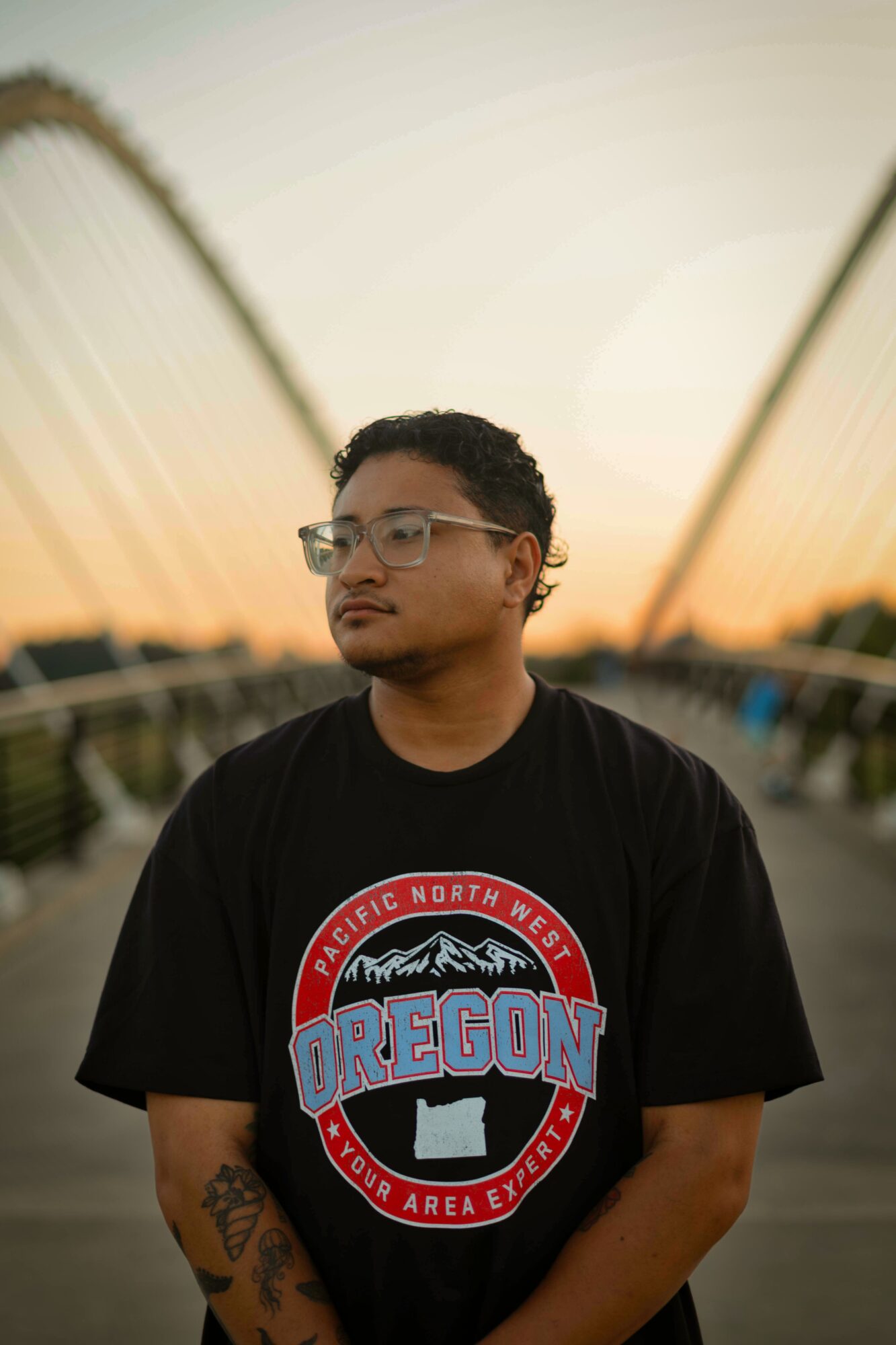 Young man with glasses and tattoos stands outdoors during sunset, wearing a black Oregon t-shirt, with a bridge in the background.
