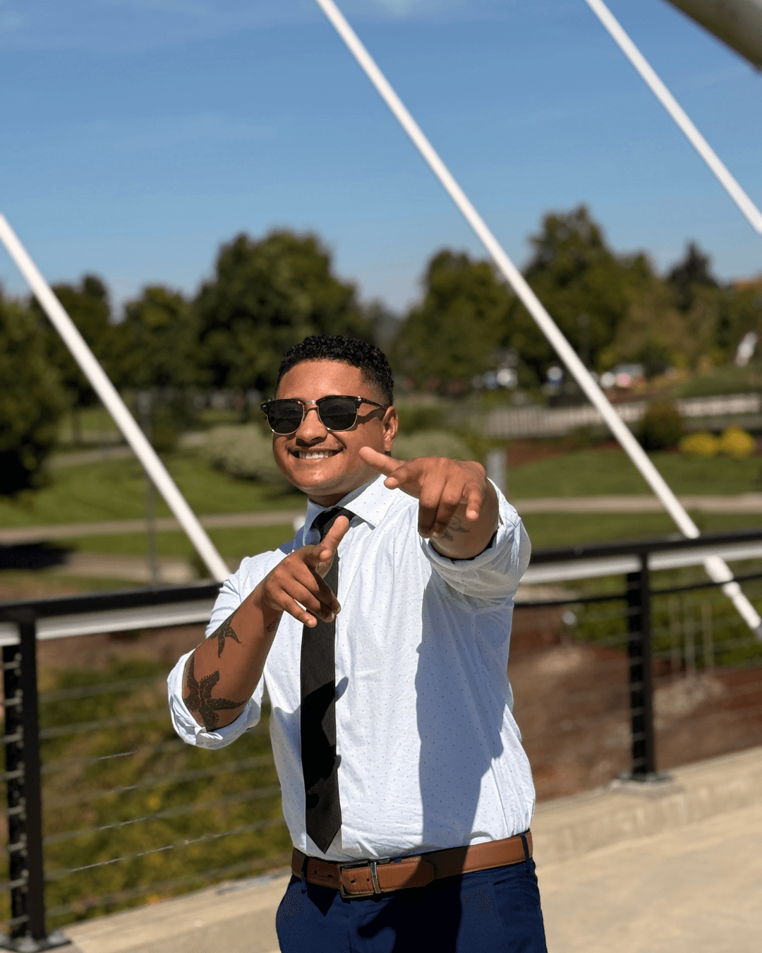 Smiling man in sunglasses, white shirt, black tie, pointing at camera outdoors with bridge and trees in background.