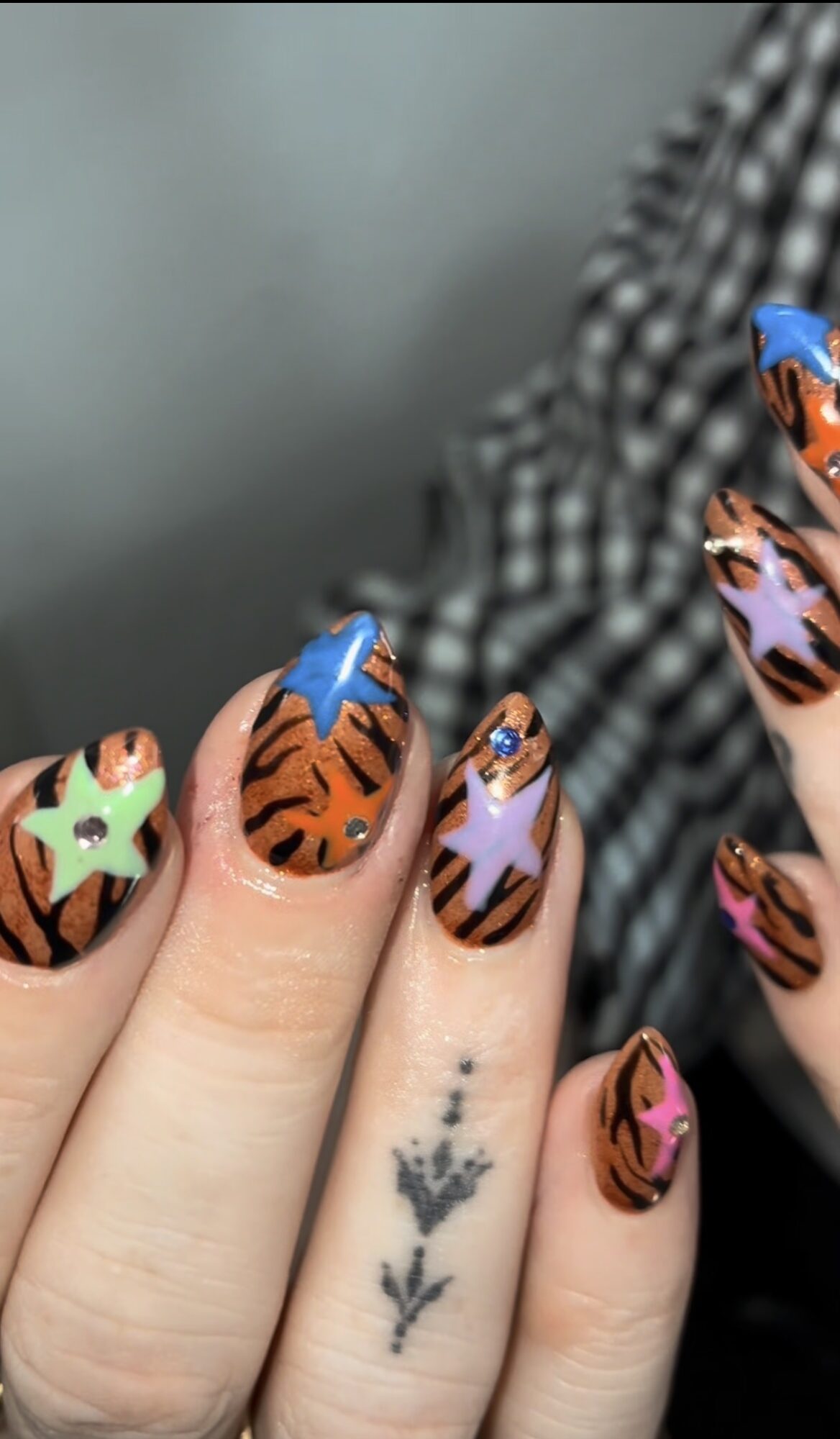 Close-up of a hand with colorful animal-themed nail art, including tiger, zebra, and leopard patterns, and a small black tattoo of a plant.