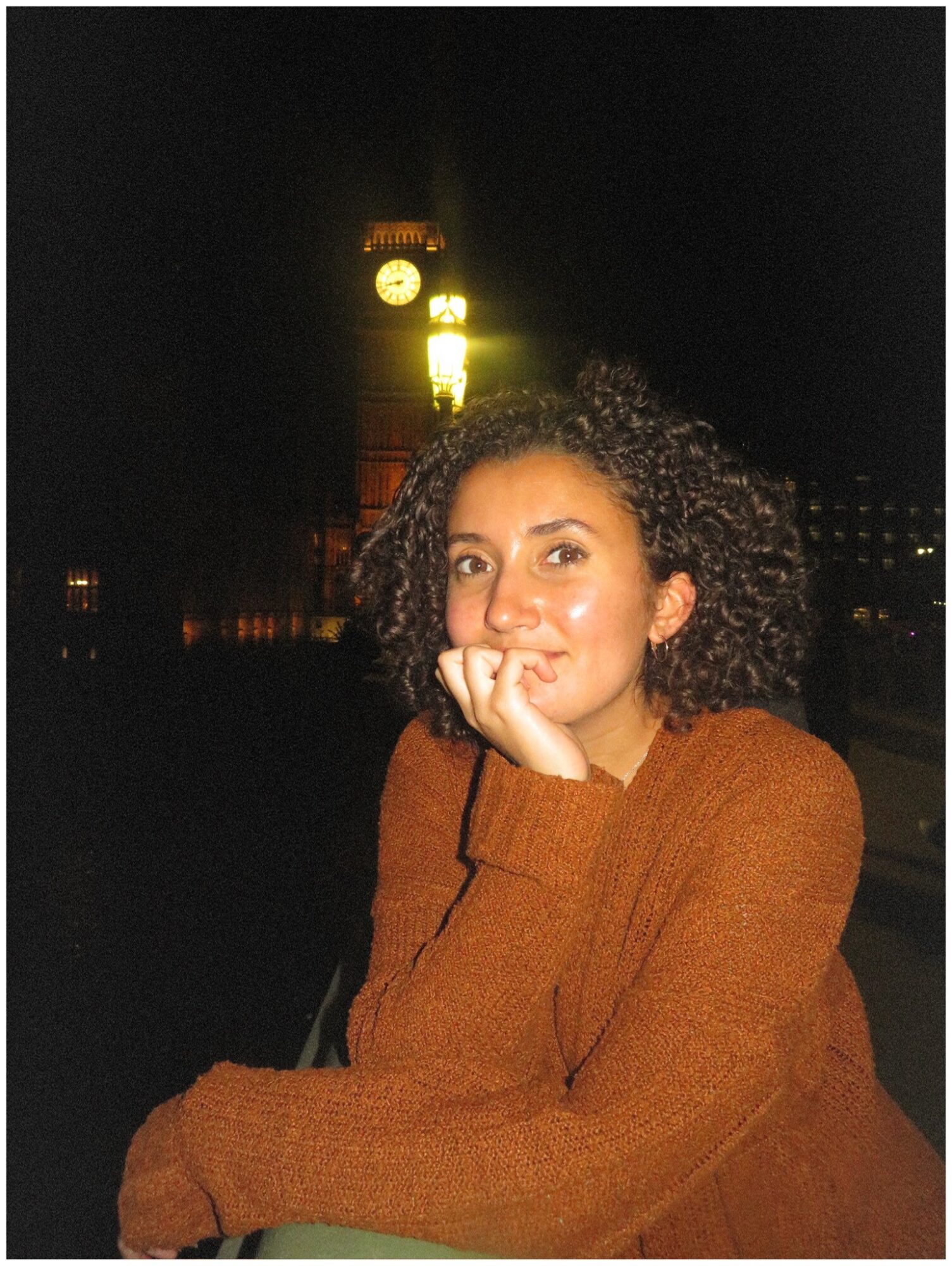 Young woman with curly hair wearing a brown sweater, resting her chin on her hand outdoors at night, illuminated clock tower in background.