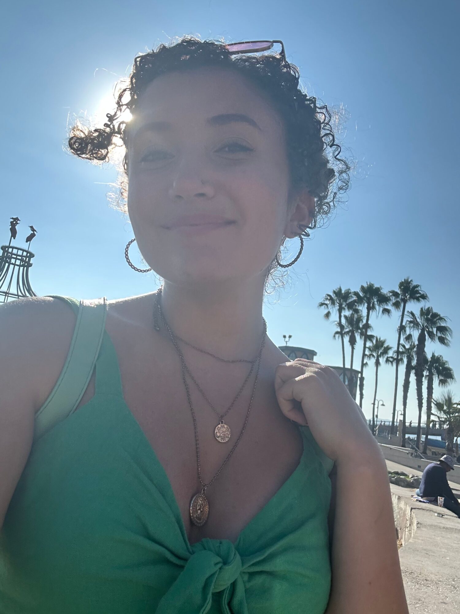 Woman with curly hair in a green top outdoors, palm trees and a clear sky in background.