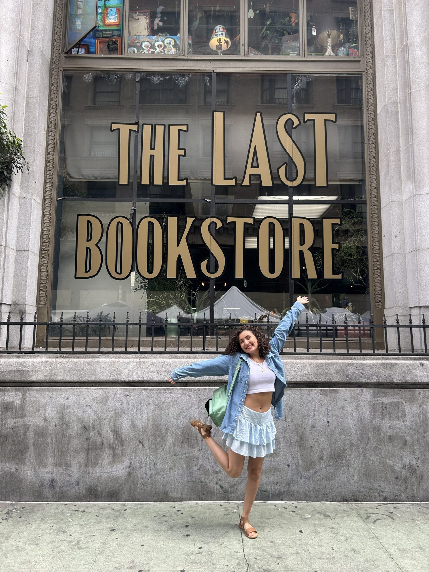 Young woman in a blue jacket and skirt poses in front of a bookstore window with gold lettering, one arm raised.
